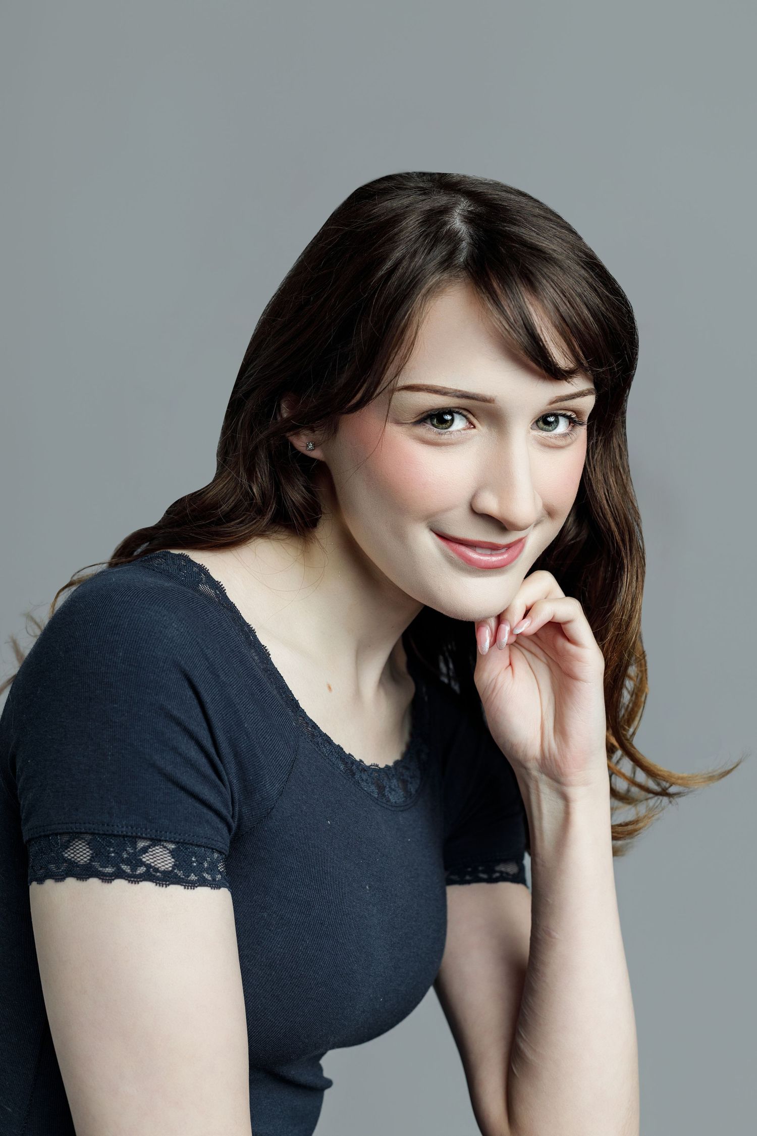 Young woman with long brown hair in a dark blue lace-trim top, smiling softly with hand near chin on gray background.