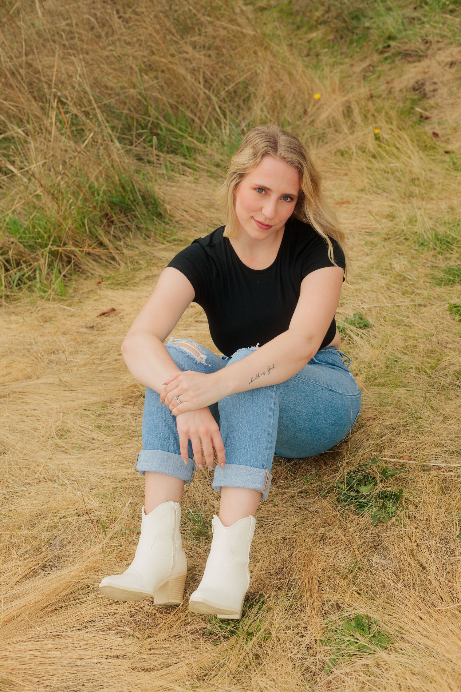 Person in black shirt and jeans sitting in dry grass field wearing white boots.