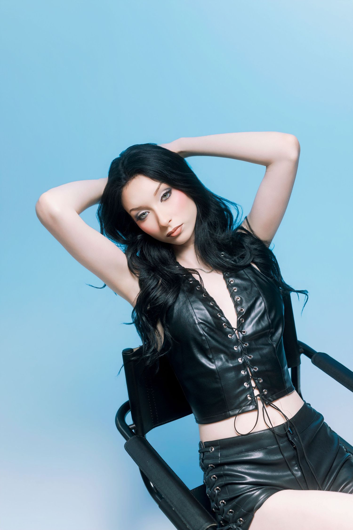 Woman in black leather corset top and skirt posing in chair against blue background with long dark hair.