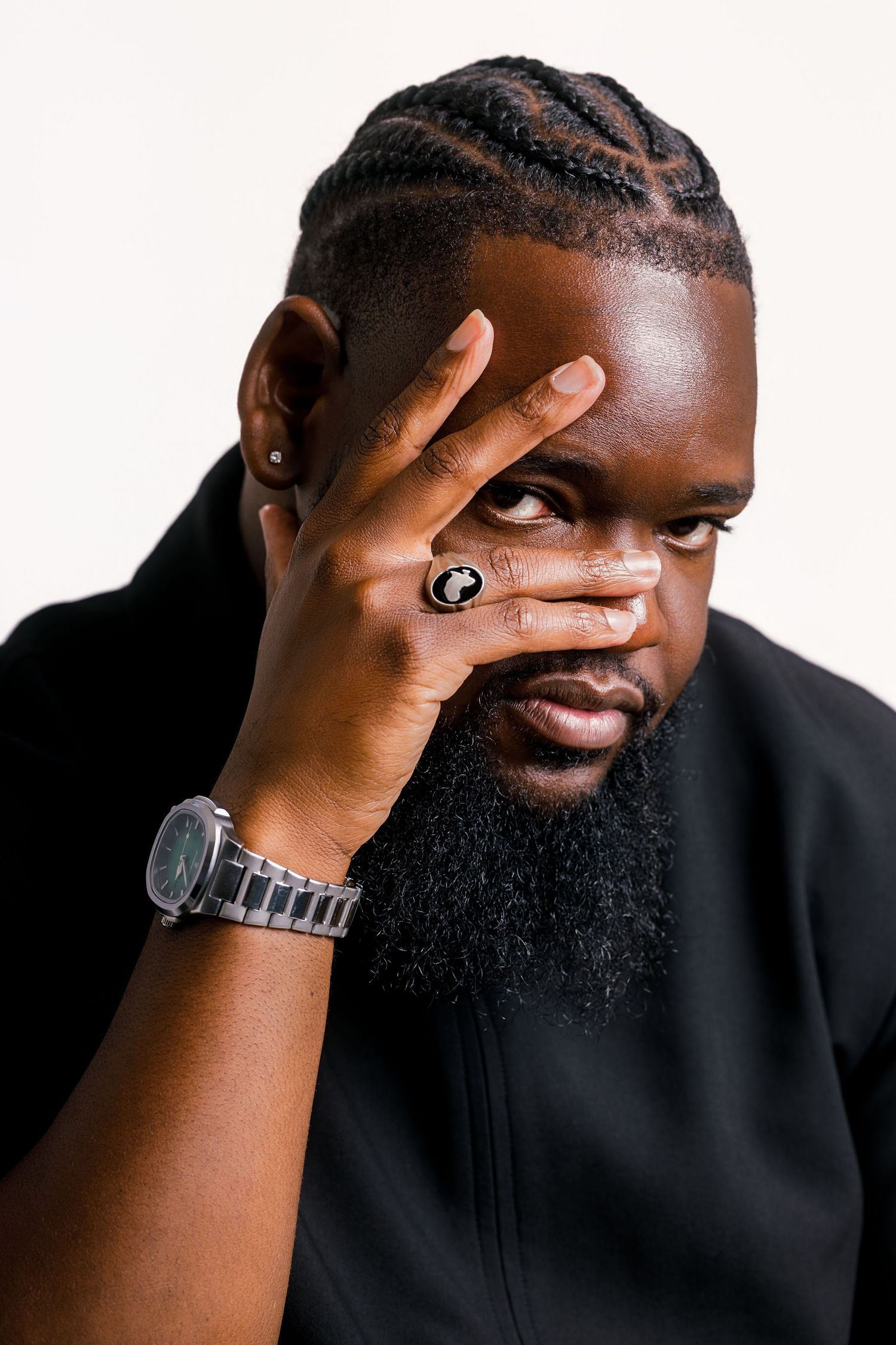 Black man with braided hair and beard covers face with hand, wearing a silver watch and ring, against white background.