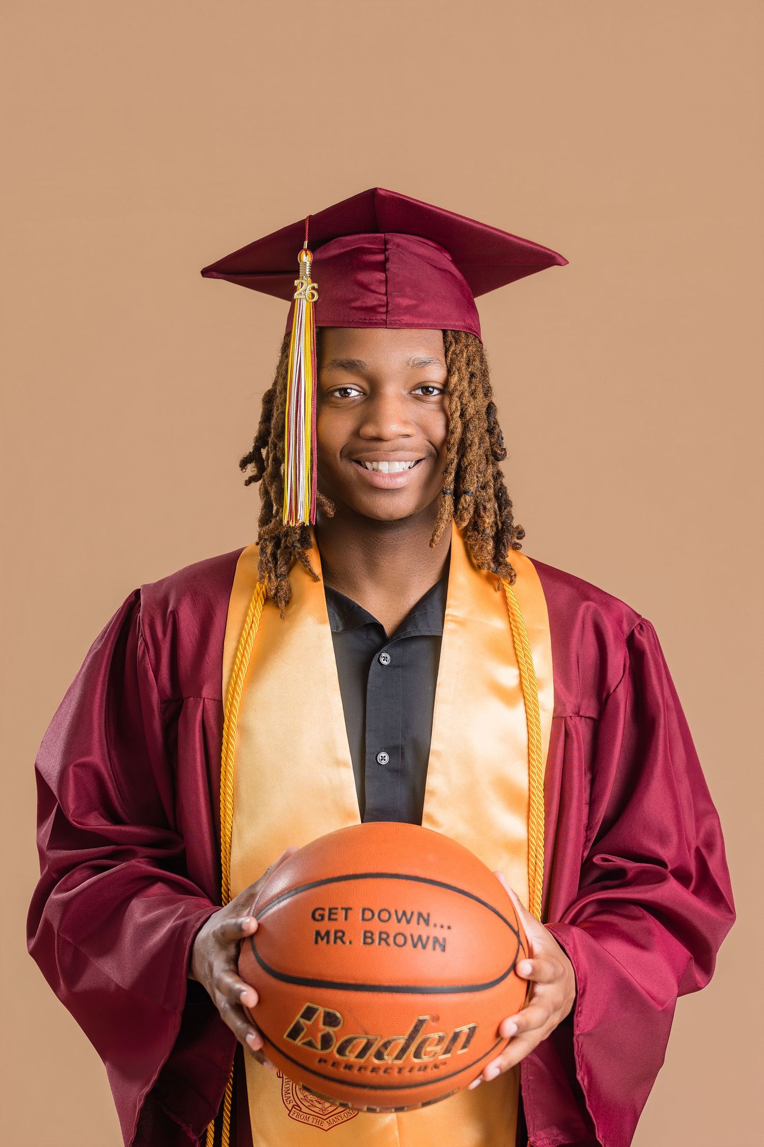Young man in maroon graduation cap and gown holding a basketball, smiling against a beige background.
