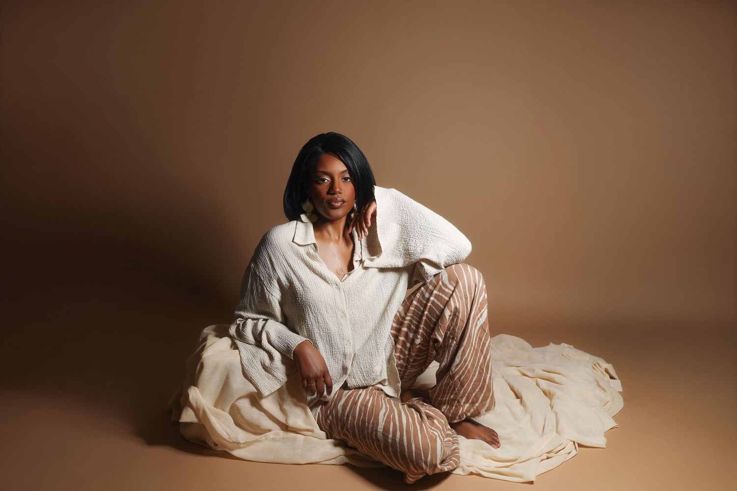 A Black woman in a white blouse and striped pants sits gracefully on the floor against a warm beige studio background.