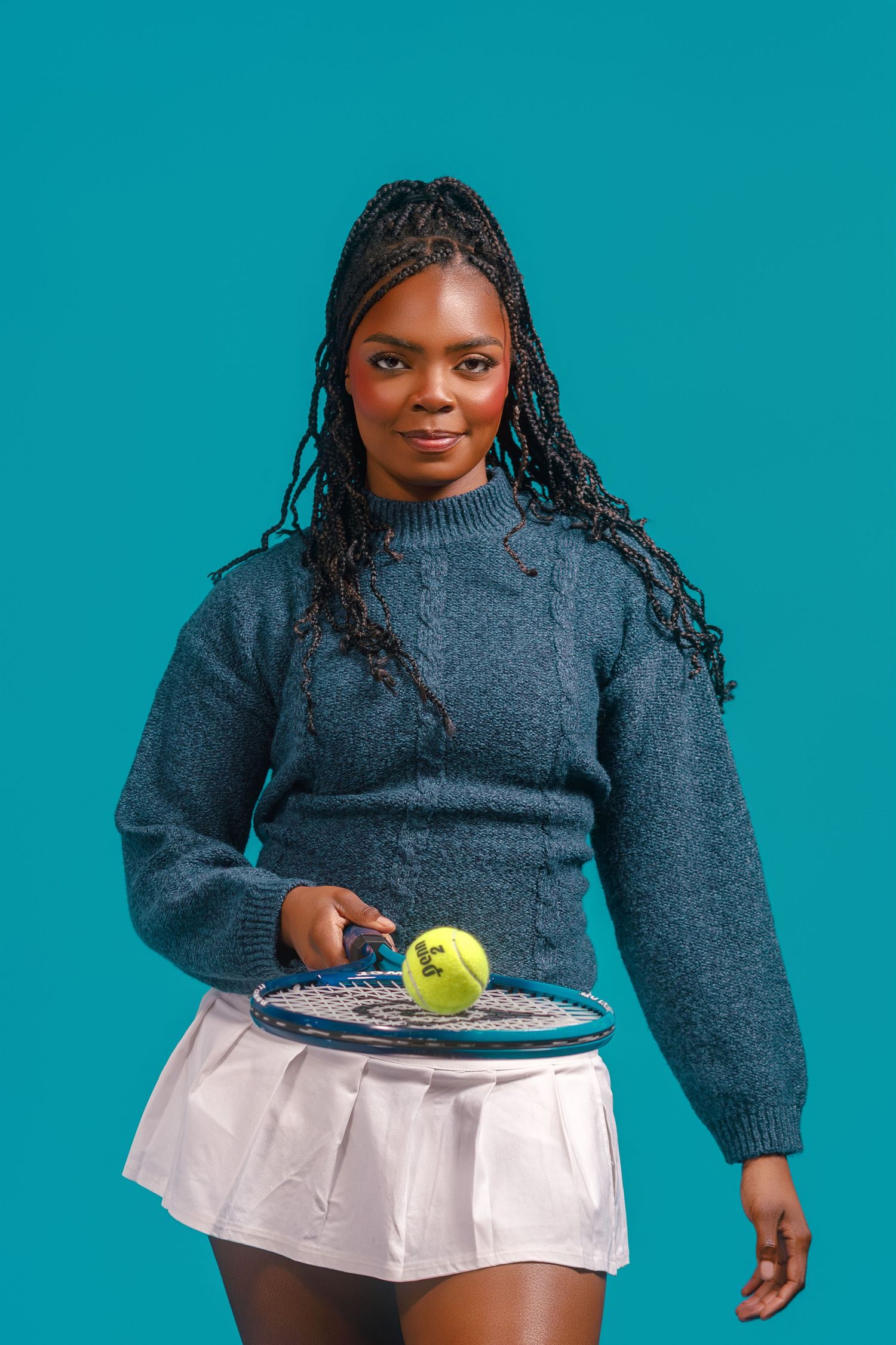 Young Black woman with braids holding a tennis racket and ball, wearing a blue sweater and white skirt on teal background.