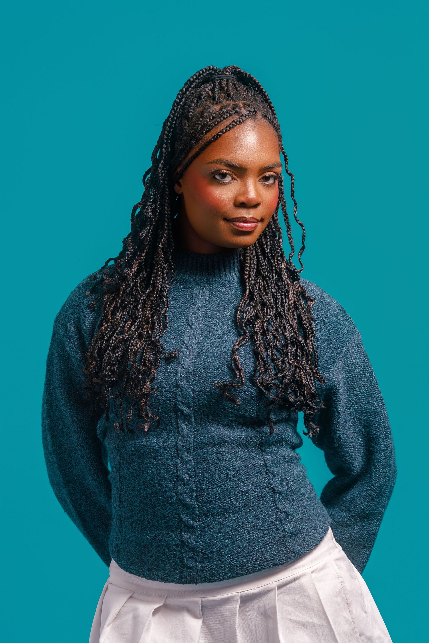 A young Black woman with long box braids wearing a teal cable-knit sweater poses against a matching teal background.