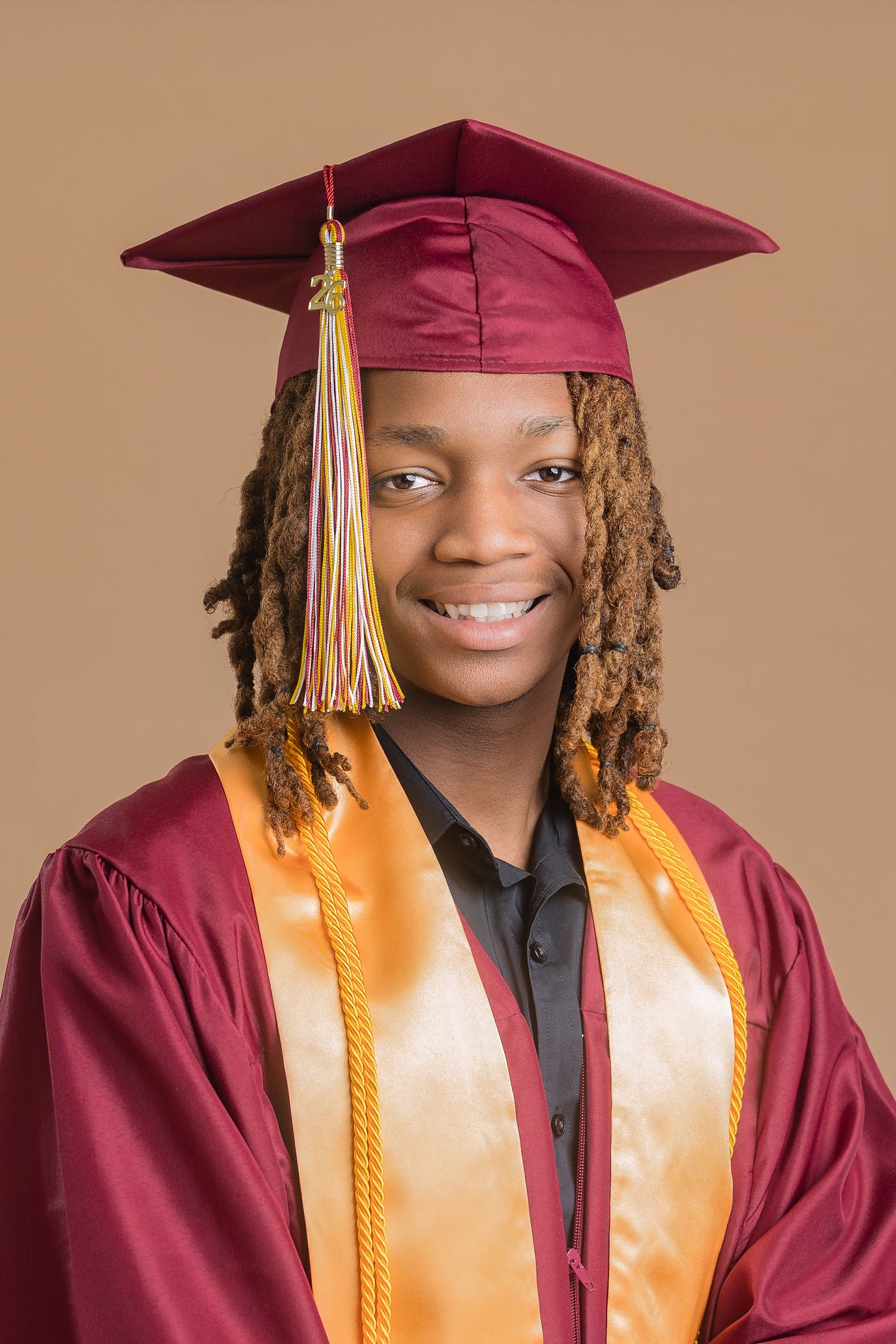 Smiling graduate in maroon cap and gown with gold honor stole, wearing dreadlocks, posed against beige background.