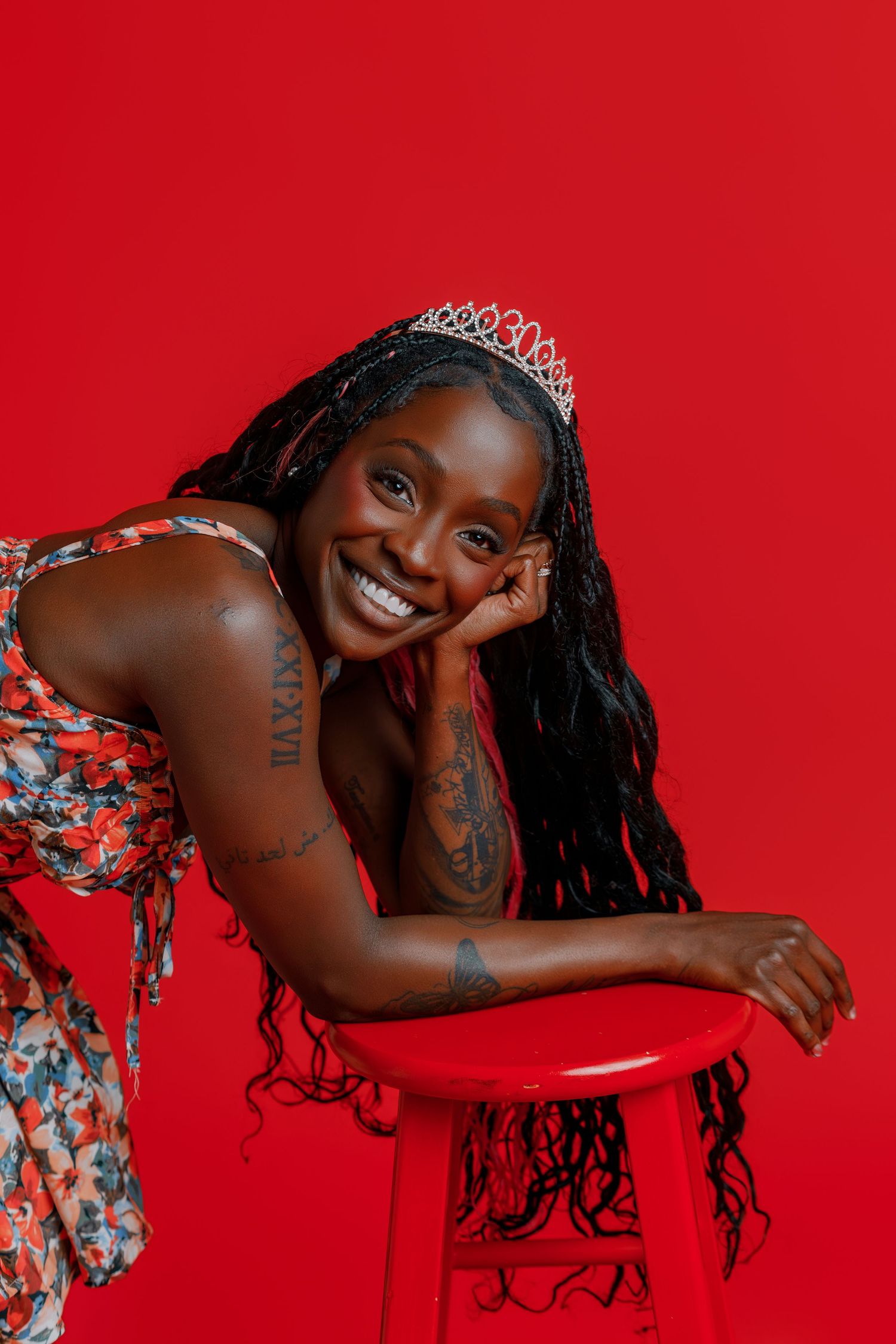 Smiling Black woman with braids, tiara, floral dress, and tattoos posing on red stool against red background.