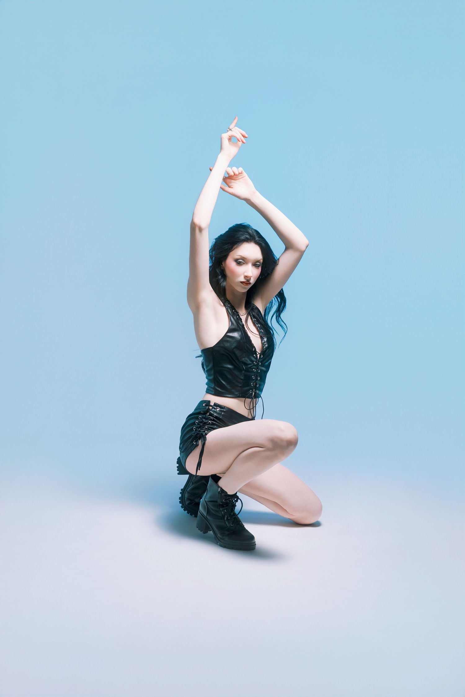 Young woman in black corset top and skirt kneeling gracefully with arms raised on a blue studio background.