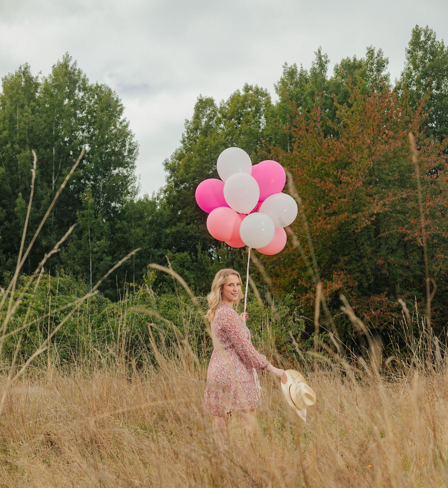 Person in a pink dress holding pink and white balloons in a tall grass field.