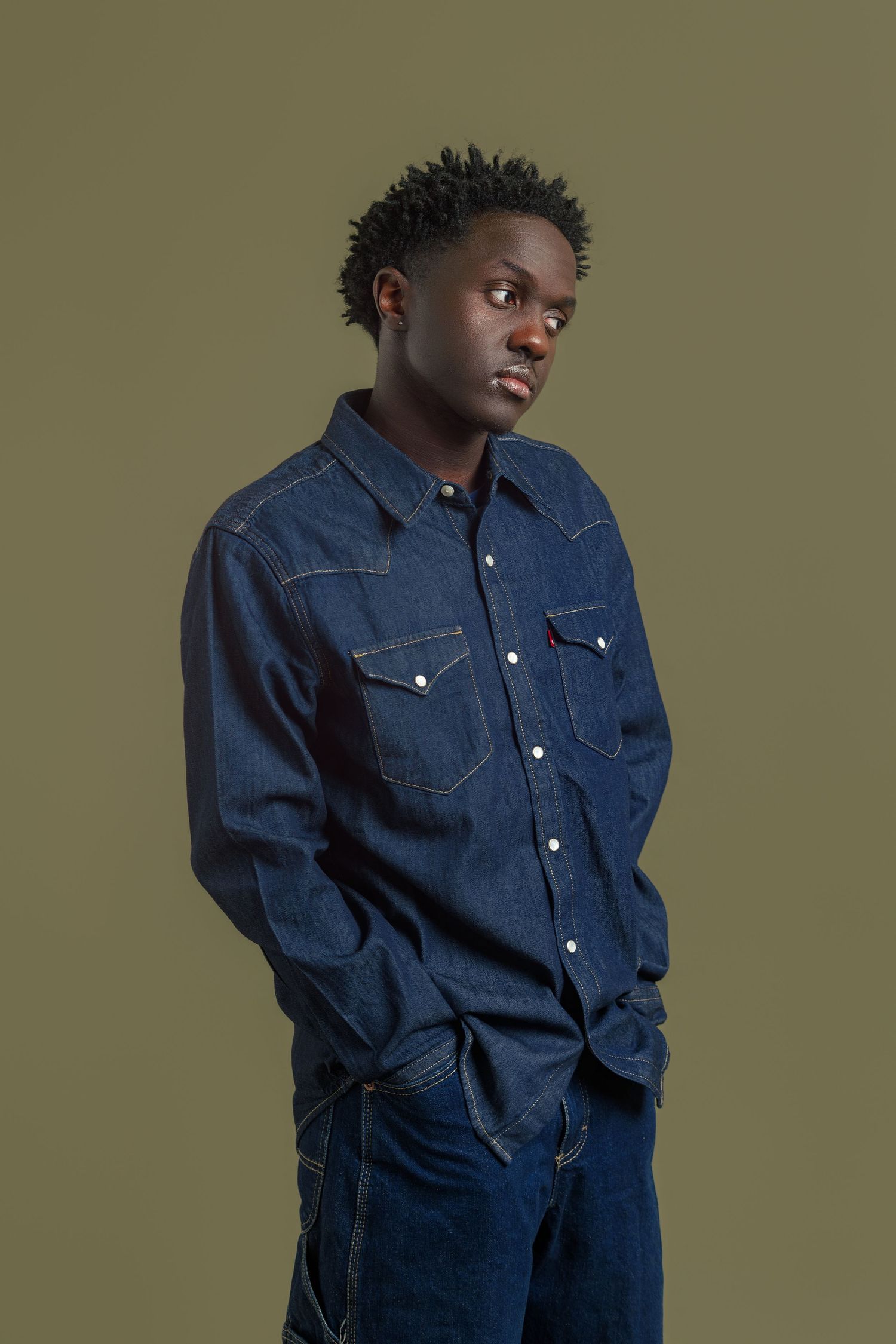 Young Black man in dark denim Levi's shirt and jeans, hands in pockets, posing against olive green background.