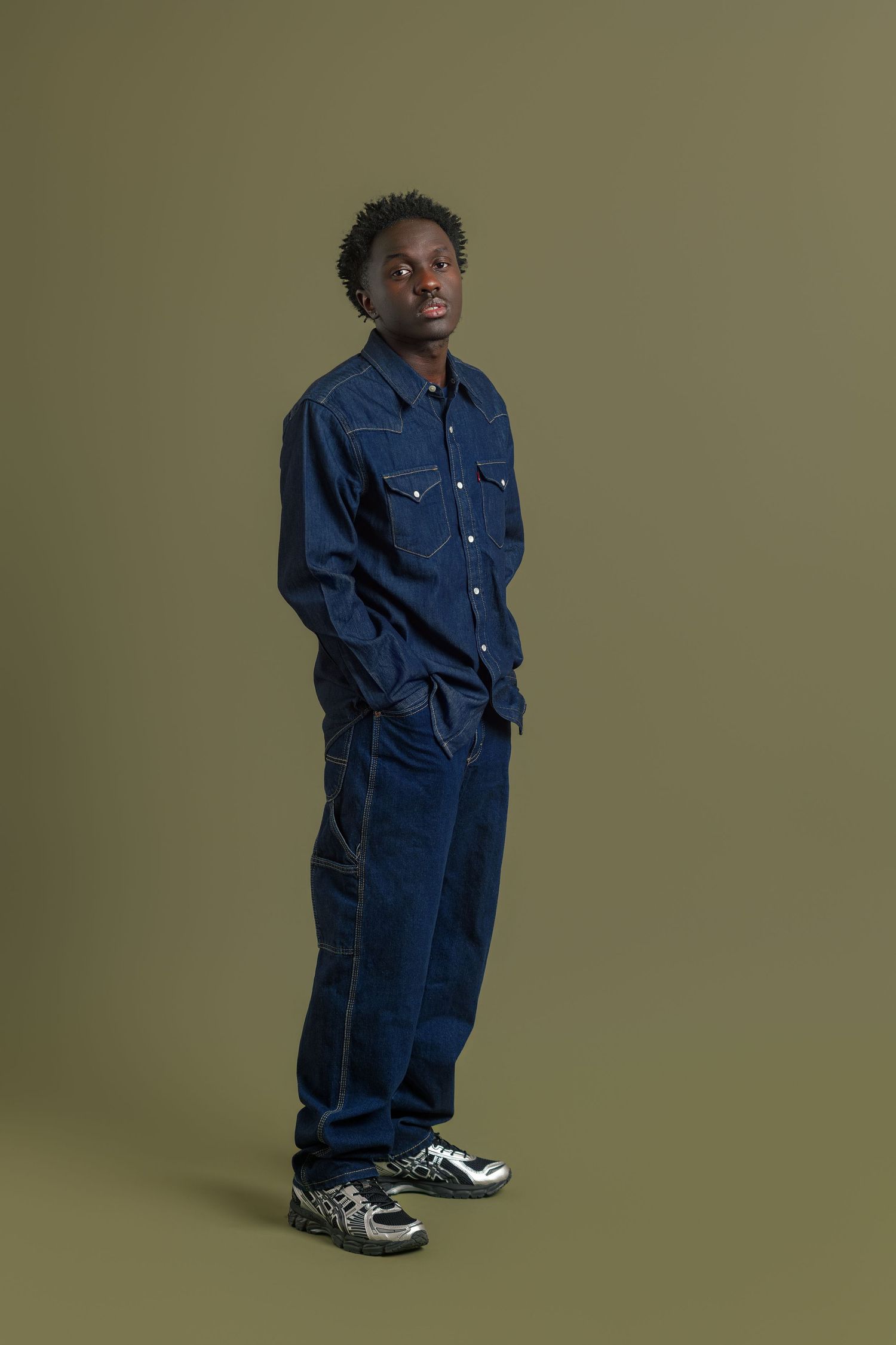 A series of fashion photos showing someone wearing an oversized navy blue denim jumpsuit with white sneakers against a green backdrop.