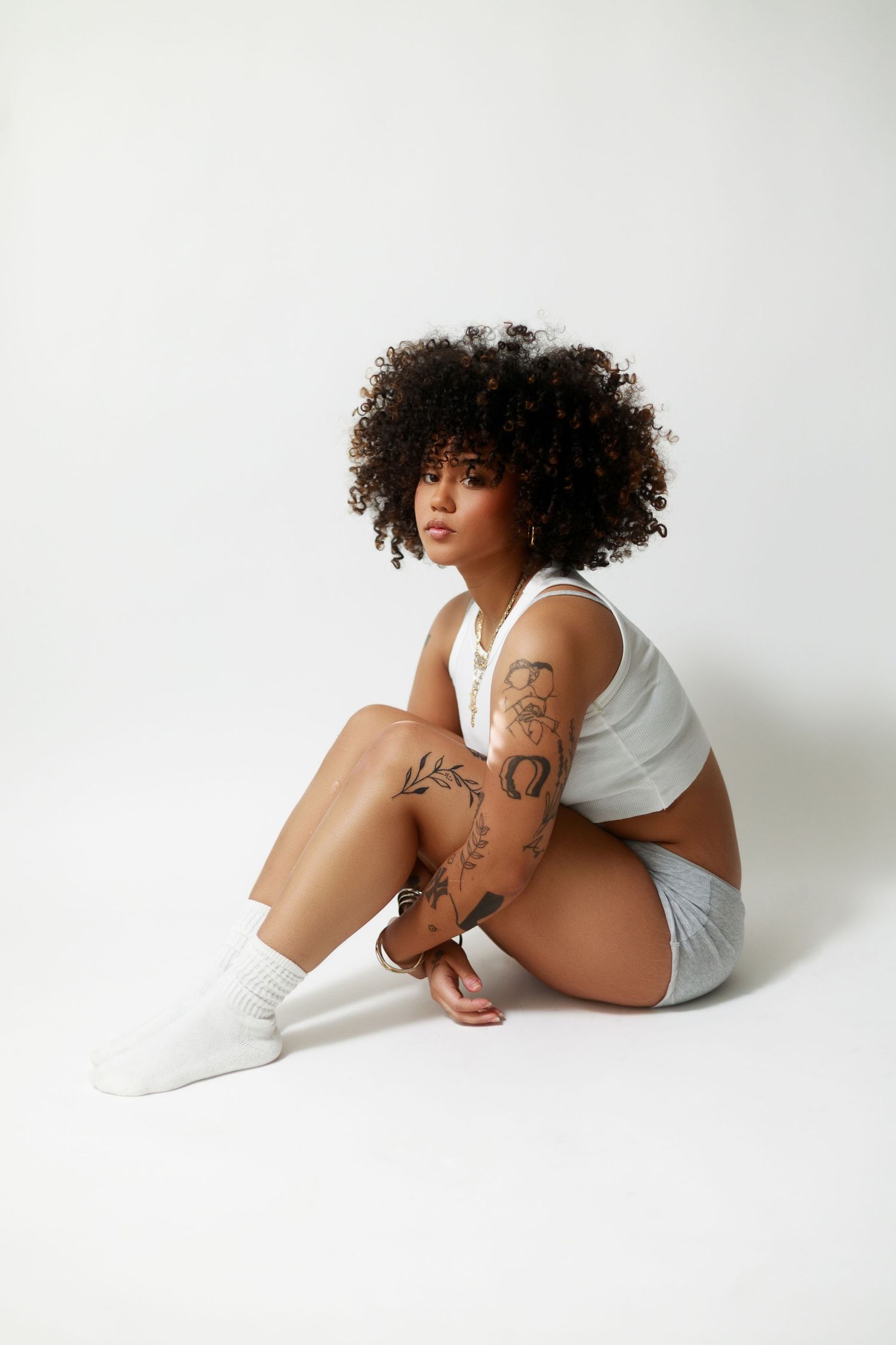Young Black woman with curly afro sits on white background wearing white shorts and crop top, showcasing floral arm tattoos.
