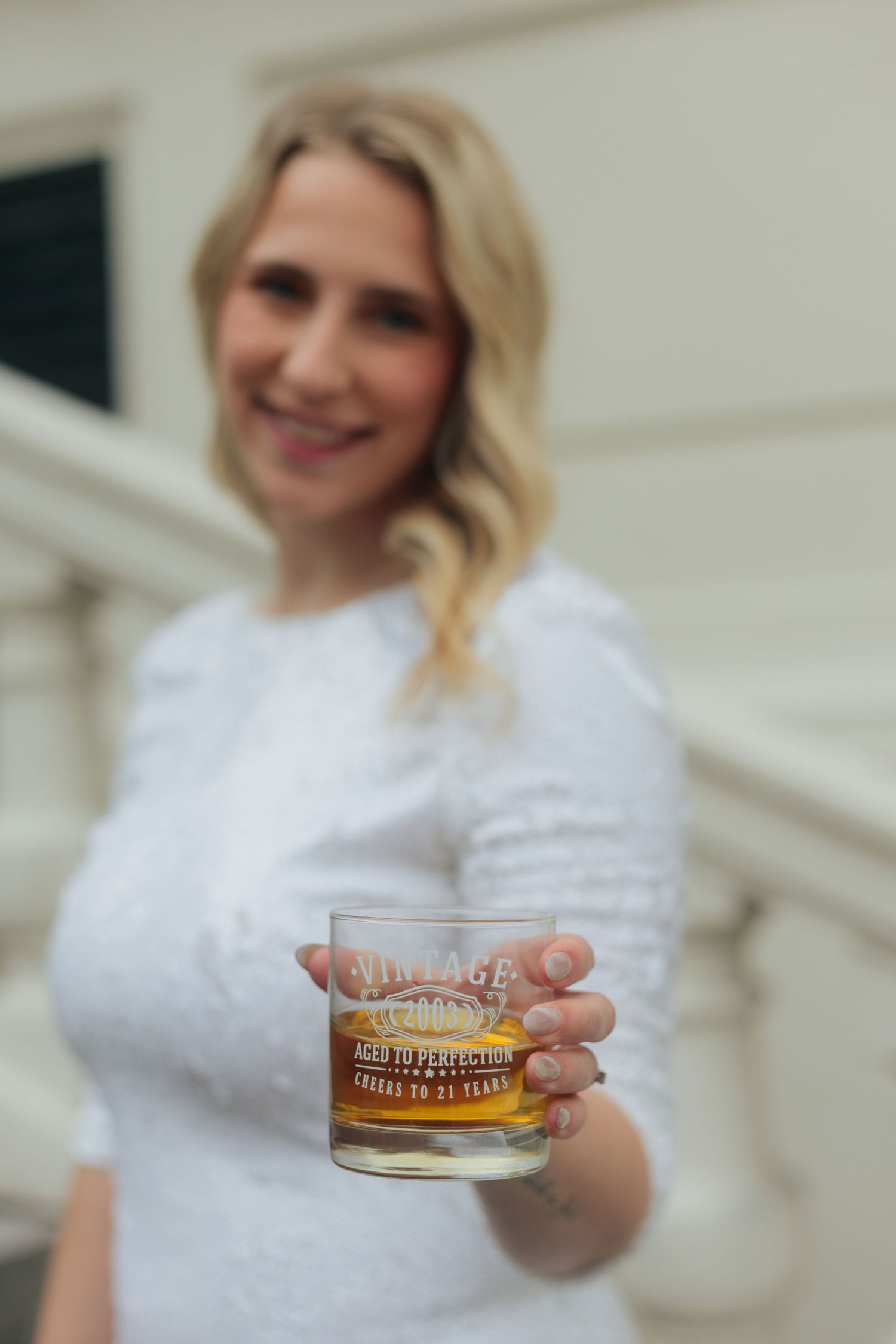 Smiling blonde woman in white dress holding a vintage whiskey glass aged to perfection, celebrating 21 years.