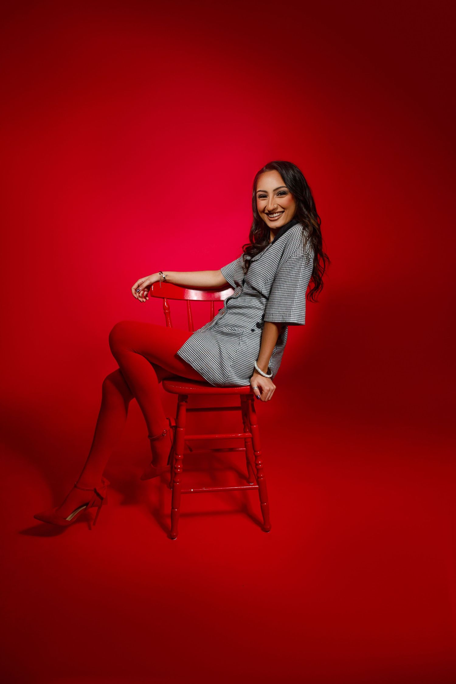 Smiling woman in gray dress and red tights sitting on a wooden stool against a vibrant red background.