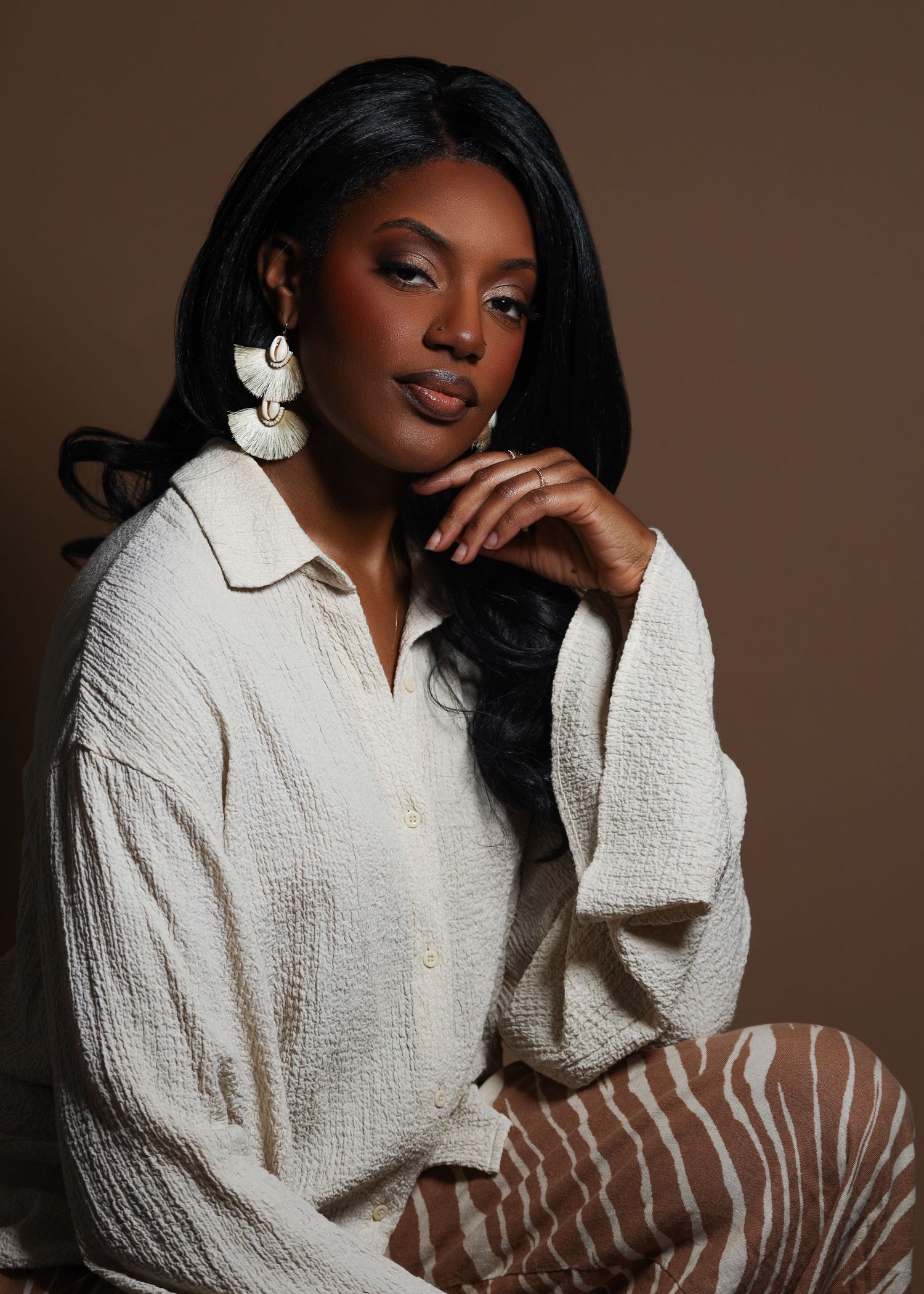 Woman in cream textured shirt and statement earrings poses gracefully against a warm brown studio background.