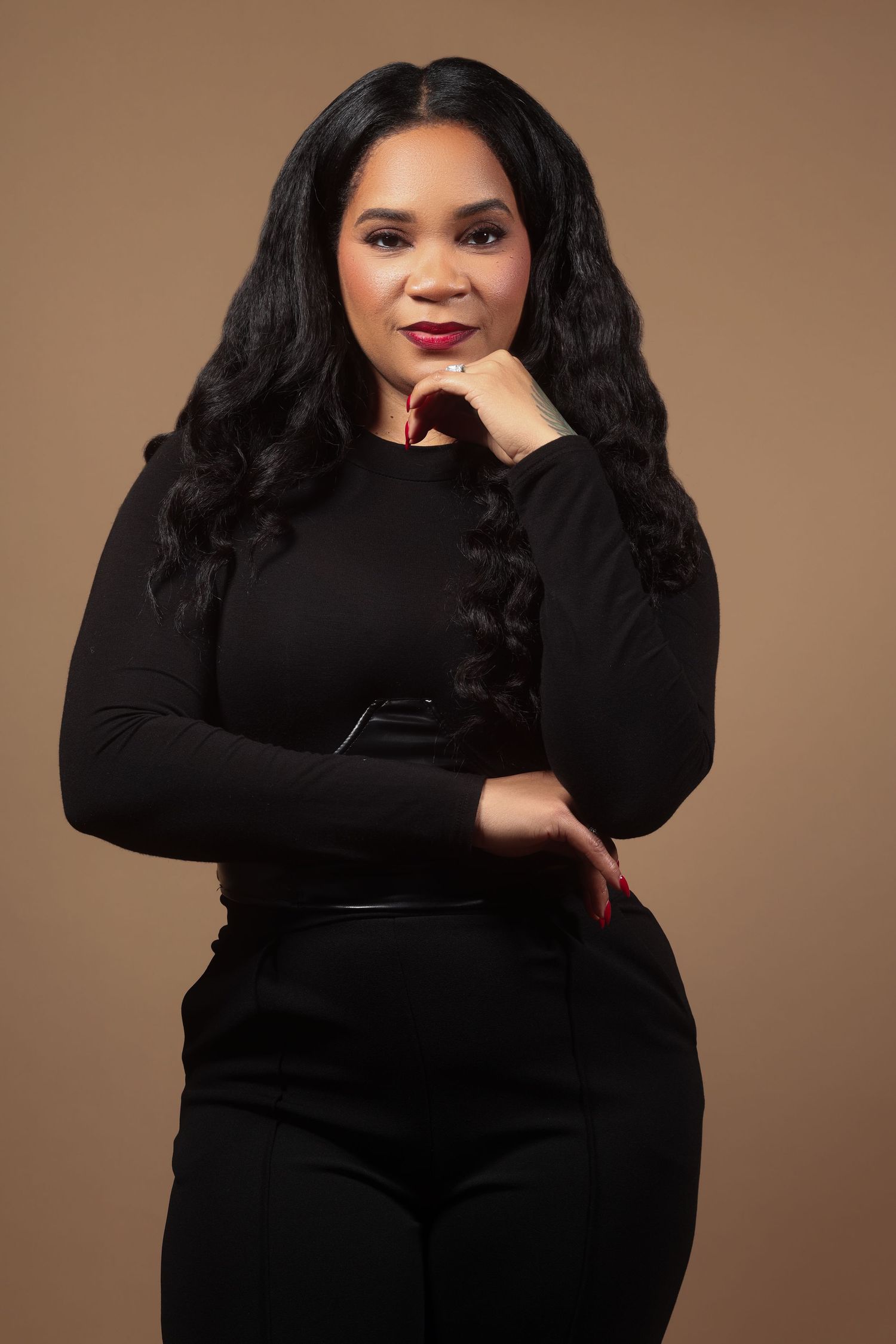 A confident woman in a black outfit poses with hand on chin against a warm brown background.