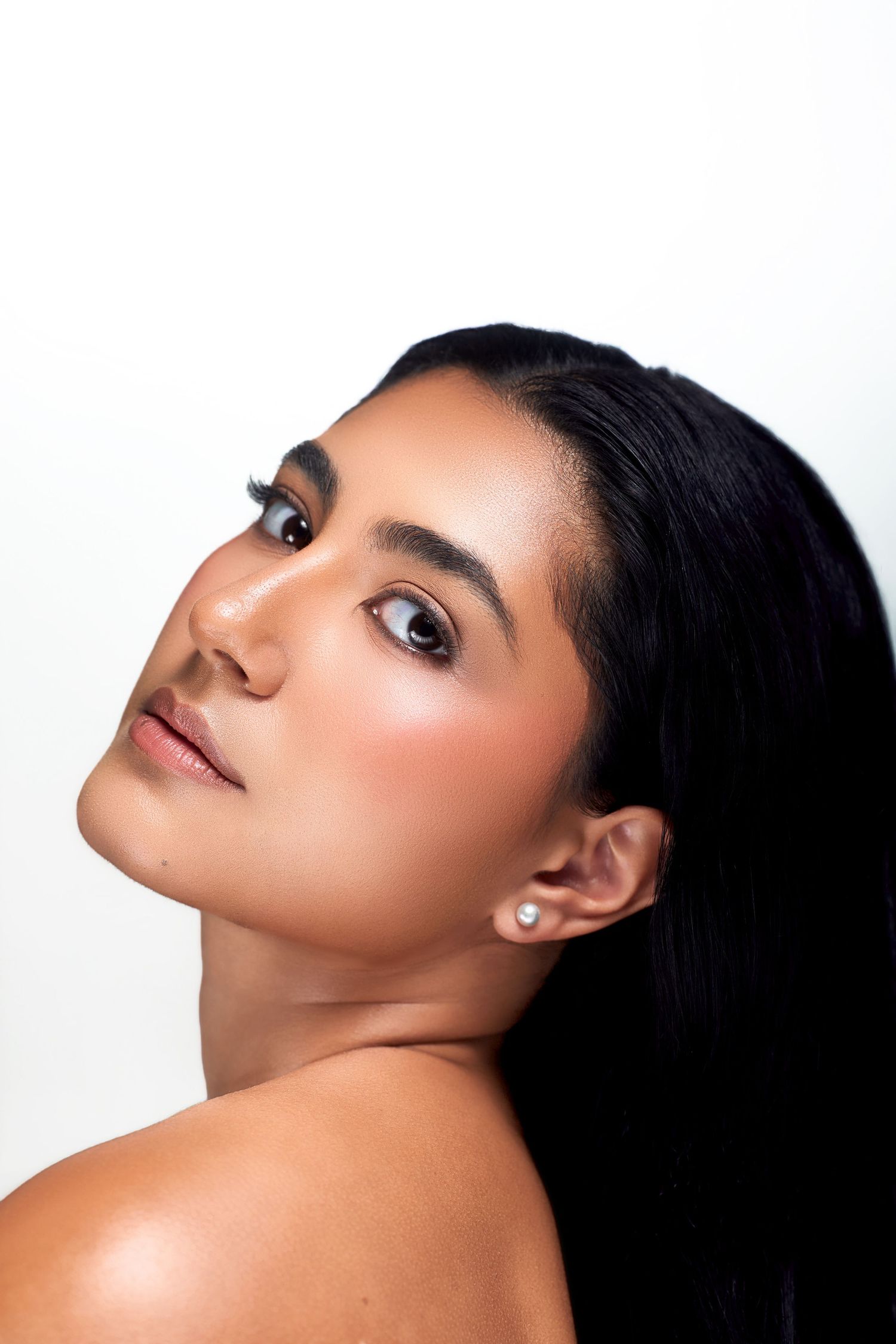 Young woman with dark hair, glowing skin, pearl earring, and rosy makeup poses against a white background.