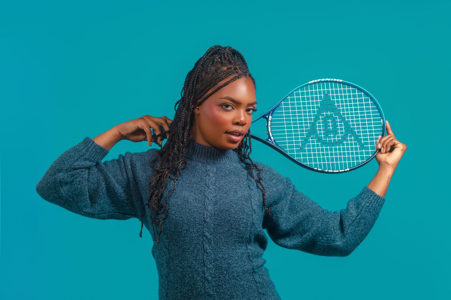 Young woman in blue sweater holding a Dunlop tennis racket against a teal background.