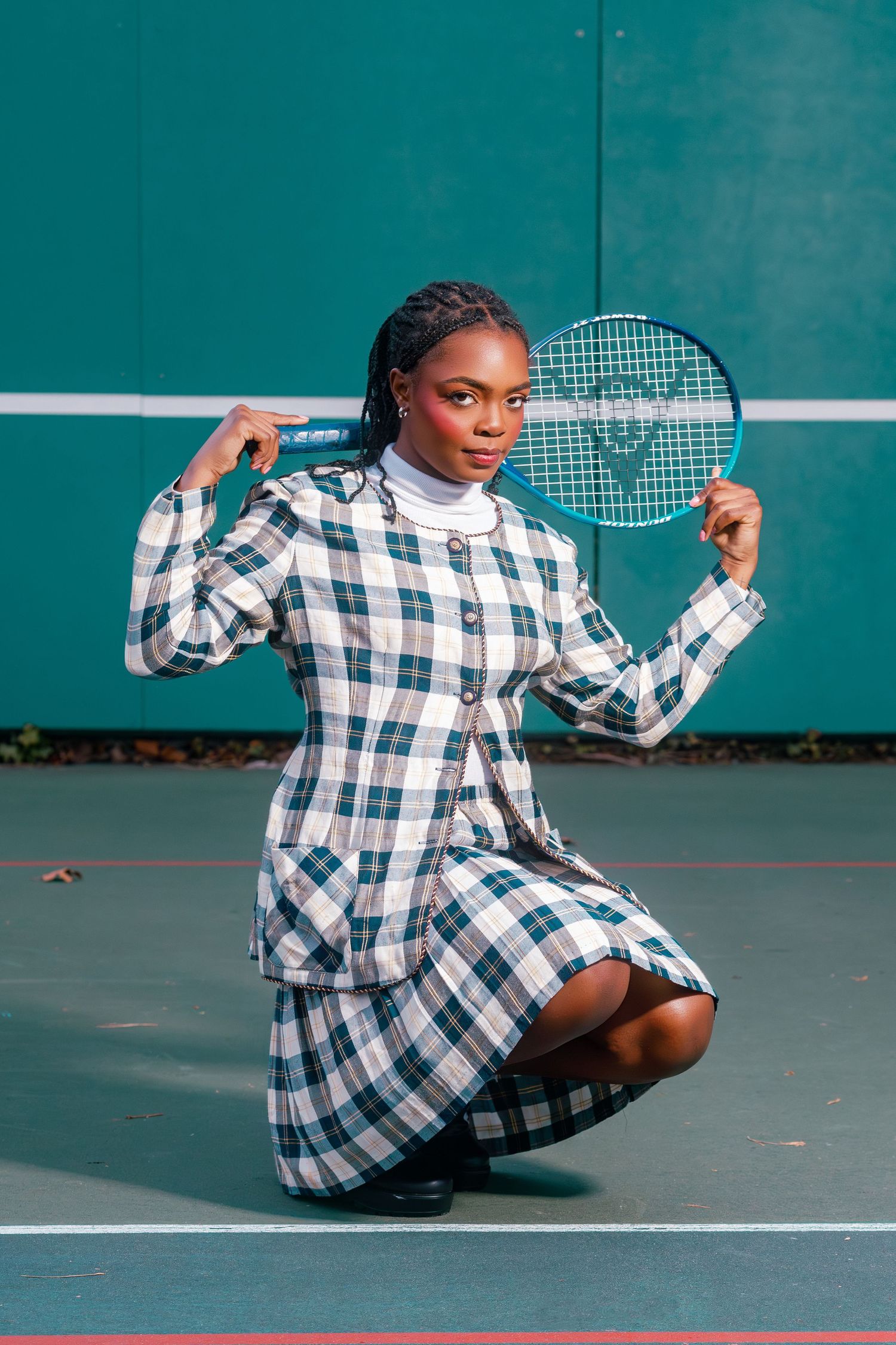 Young Black woman in plaid suit holds tennis racket behind her shoulders while posing on a teal court.