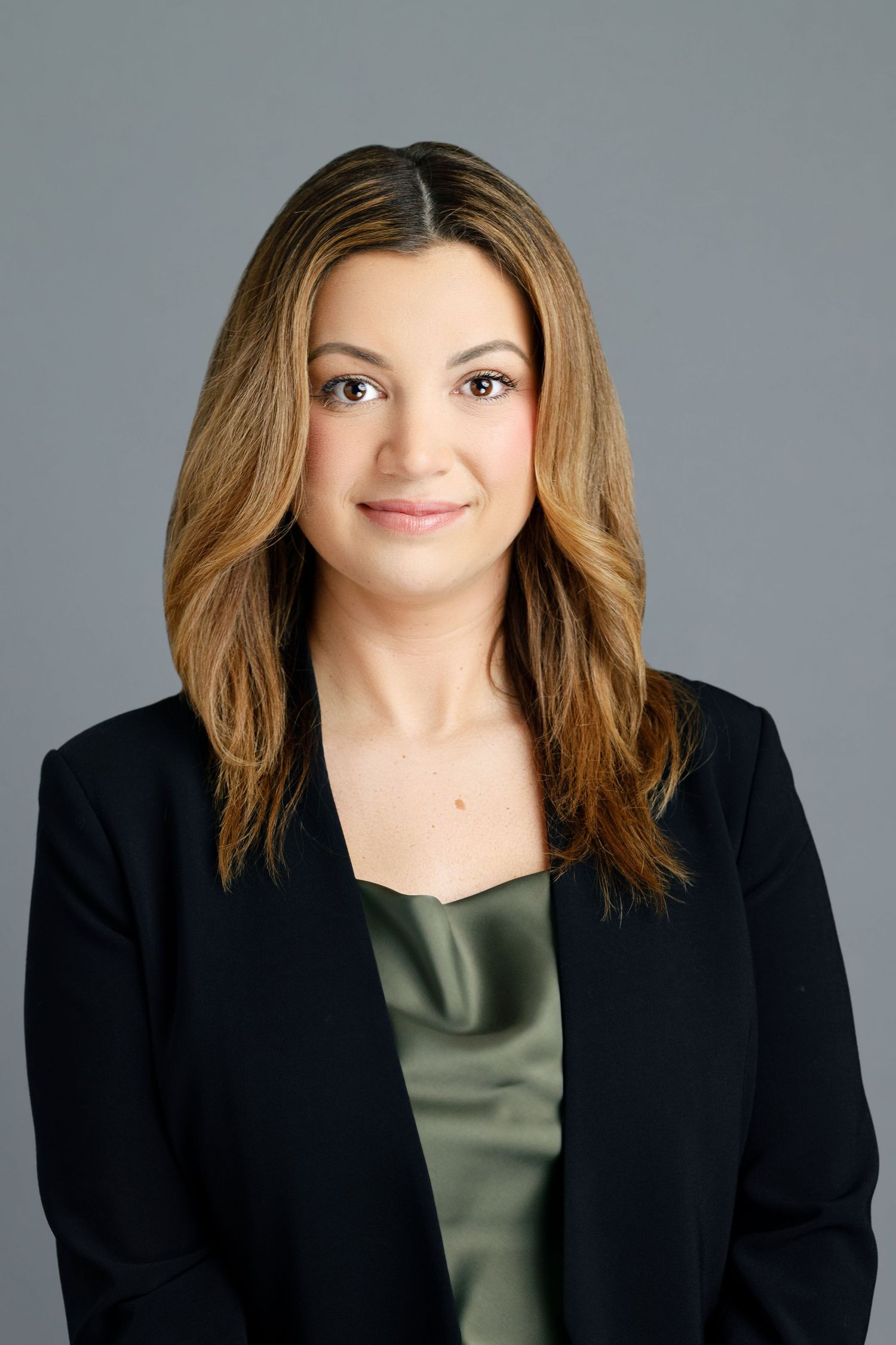 Professional headshot of a woman in a black blazer and olive satin top, smiling confidently against a gray background.