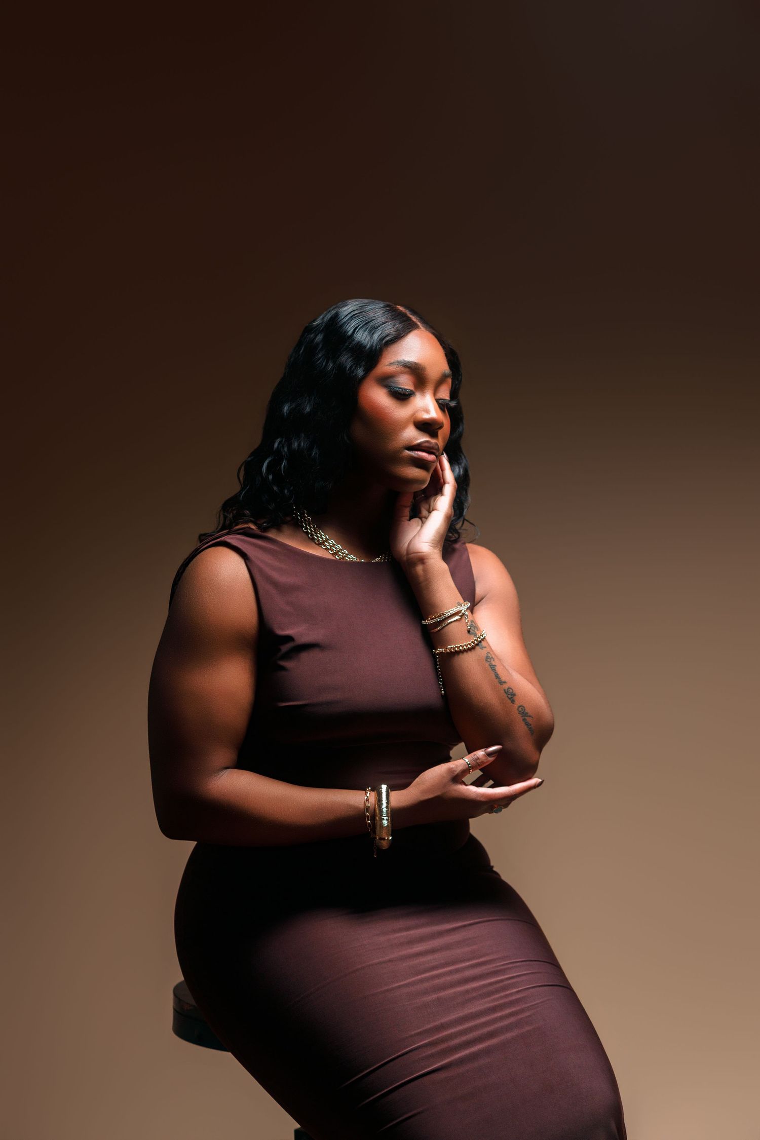 A woman in a burgundy dress poses thoughtfully with gold jewelry against a warm brown background.