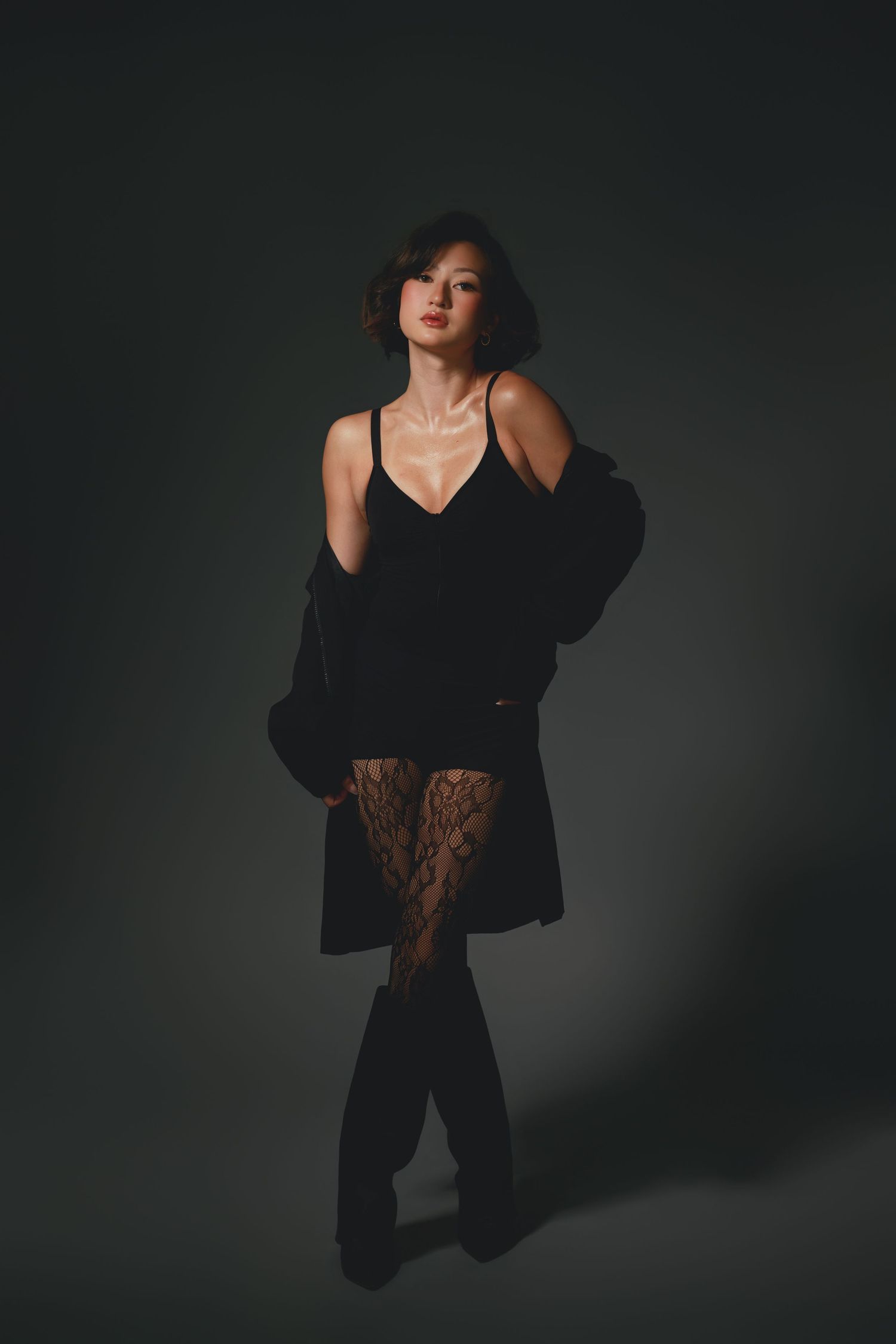 Woman in black lingerie, lace tights, and open blazer poses confidently against dark studio background.