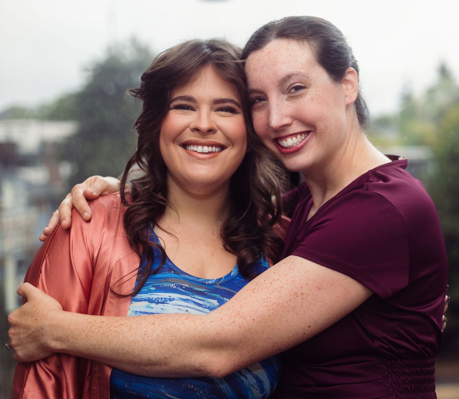 Two smiling women hugging outdoors, one in coral cardigan and blue top, other in maroon shirt, sharing a joyful embrace.