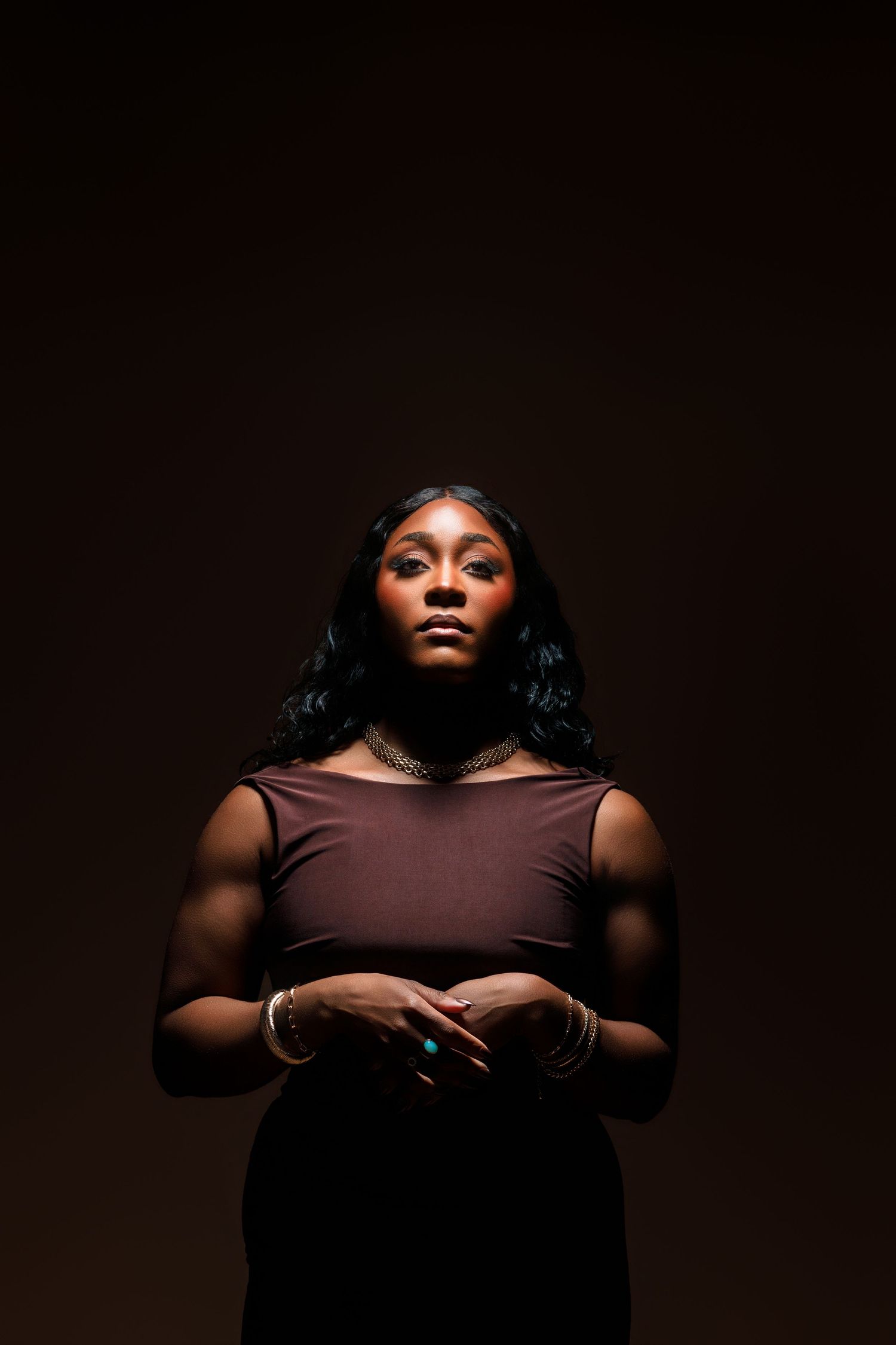 A Black woman in a brown sleeveless dress with gold jewelry poses confidently against a dark background.