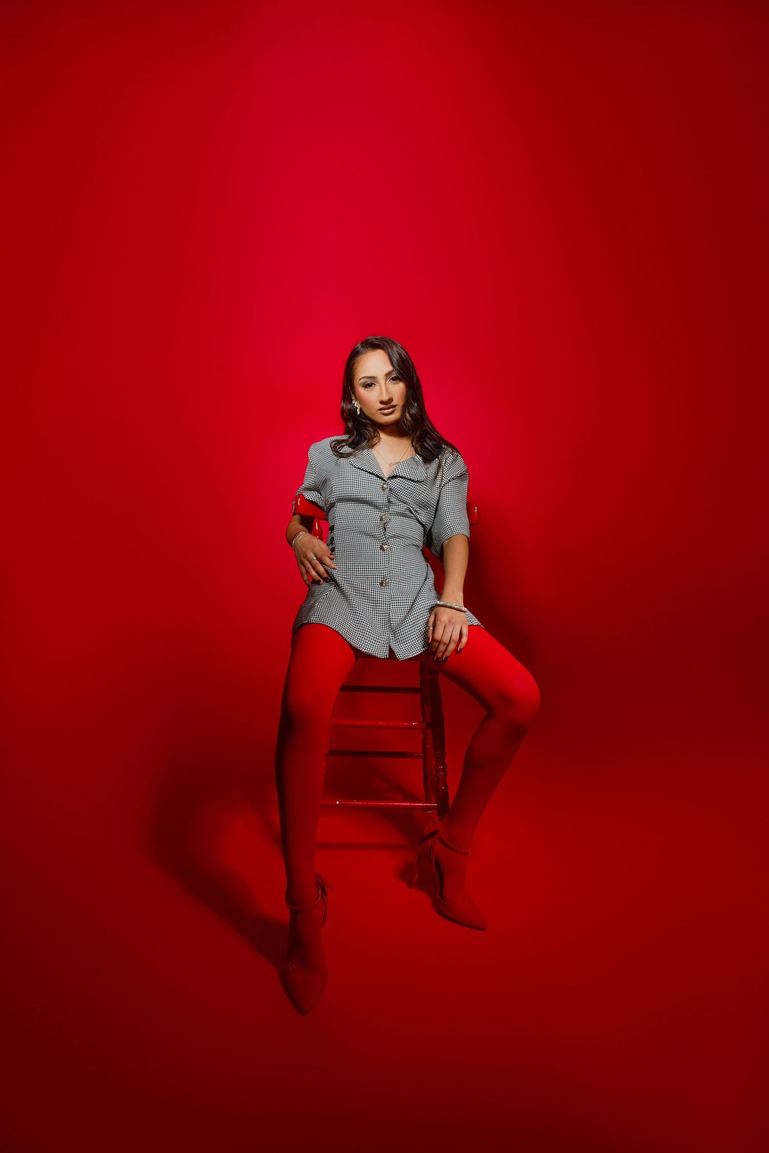 Woman in gray dress and red tights sitting on stool against vibrant red background in studio photo.