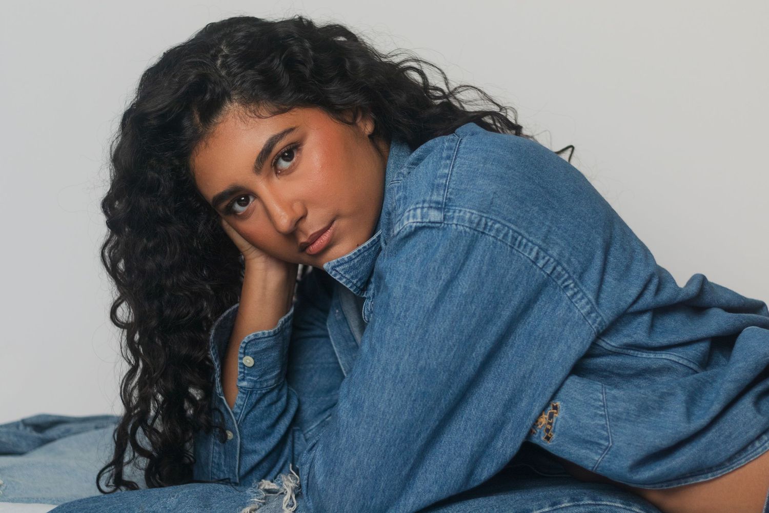 Young woman with curly dark hair wearing a denim outfit, posing gracefully against a white background.
