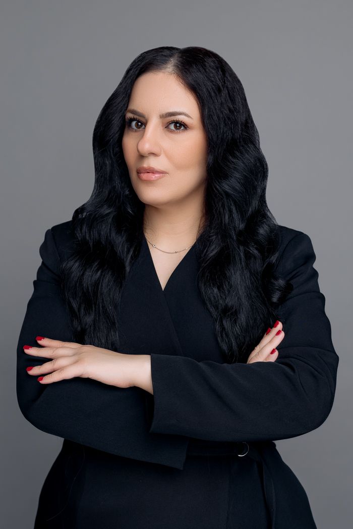 Professional woman in black blazer with crossed arms, dark wavy hair, red nails, posing confidently against gray background.