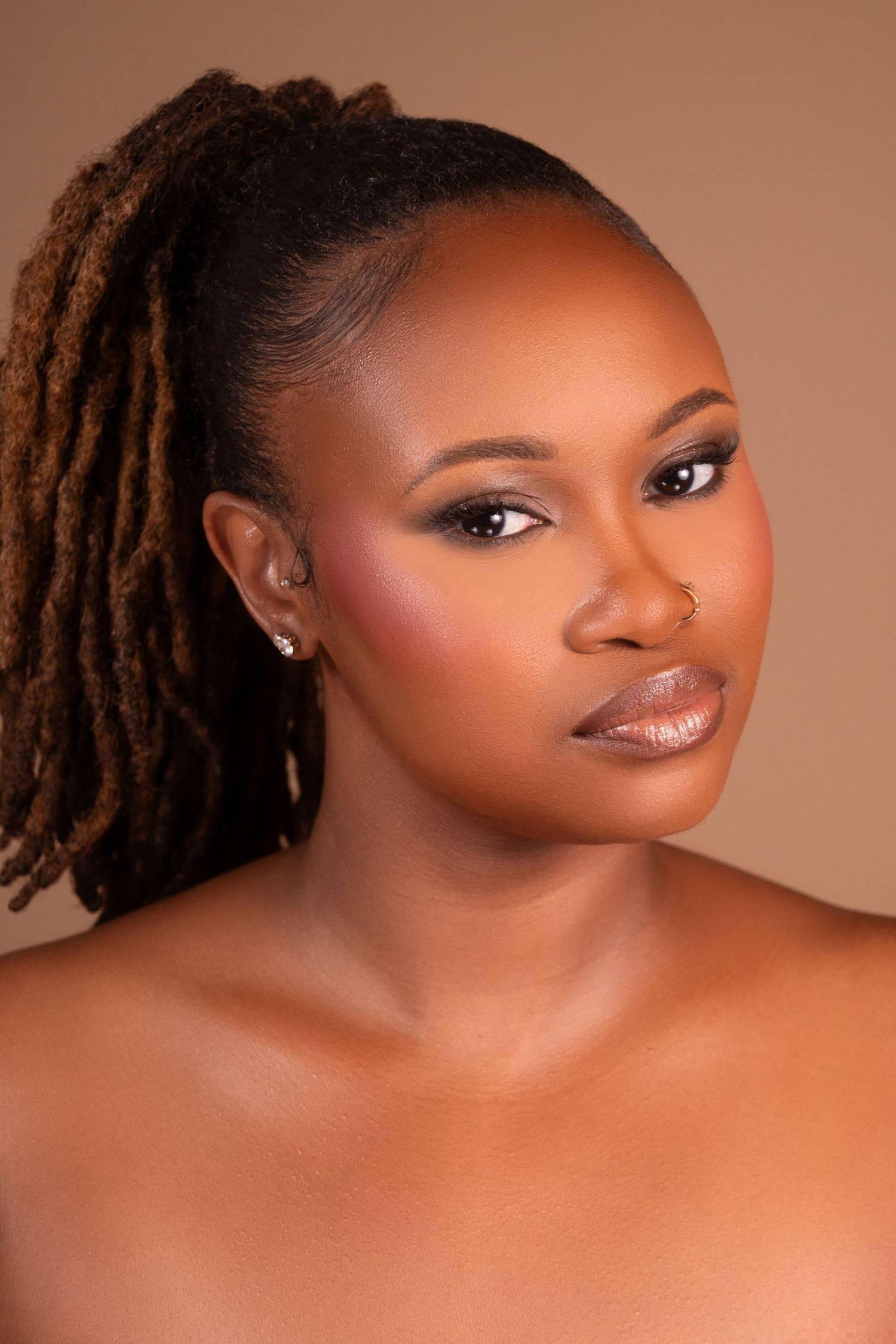 Beautiful Black woman with locs styled in a ponytail, wearing glamorous makeup and stud earrings against a beige background.