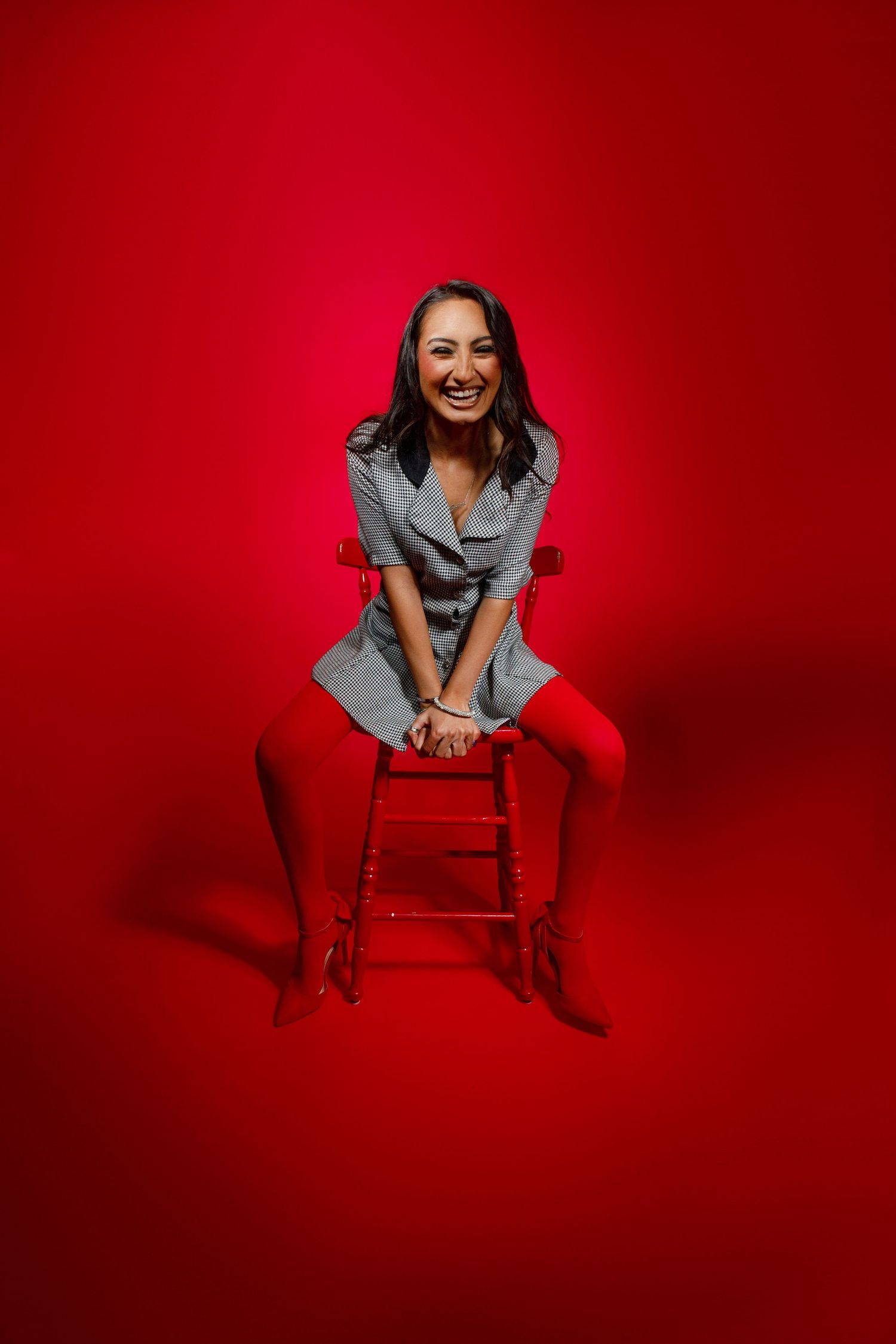 Smiling woman in gray dress and red tights sitting on wooden stool against vibrant red background.