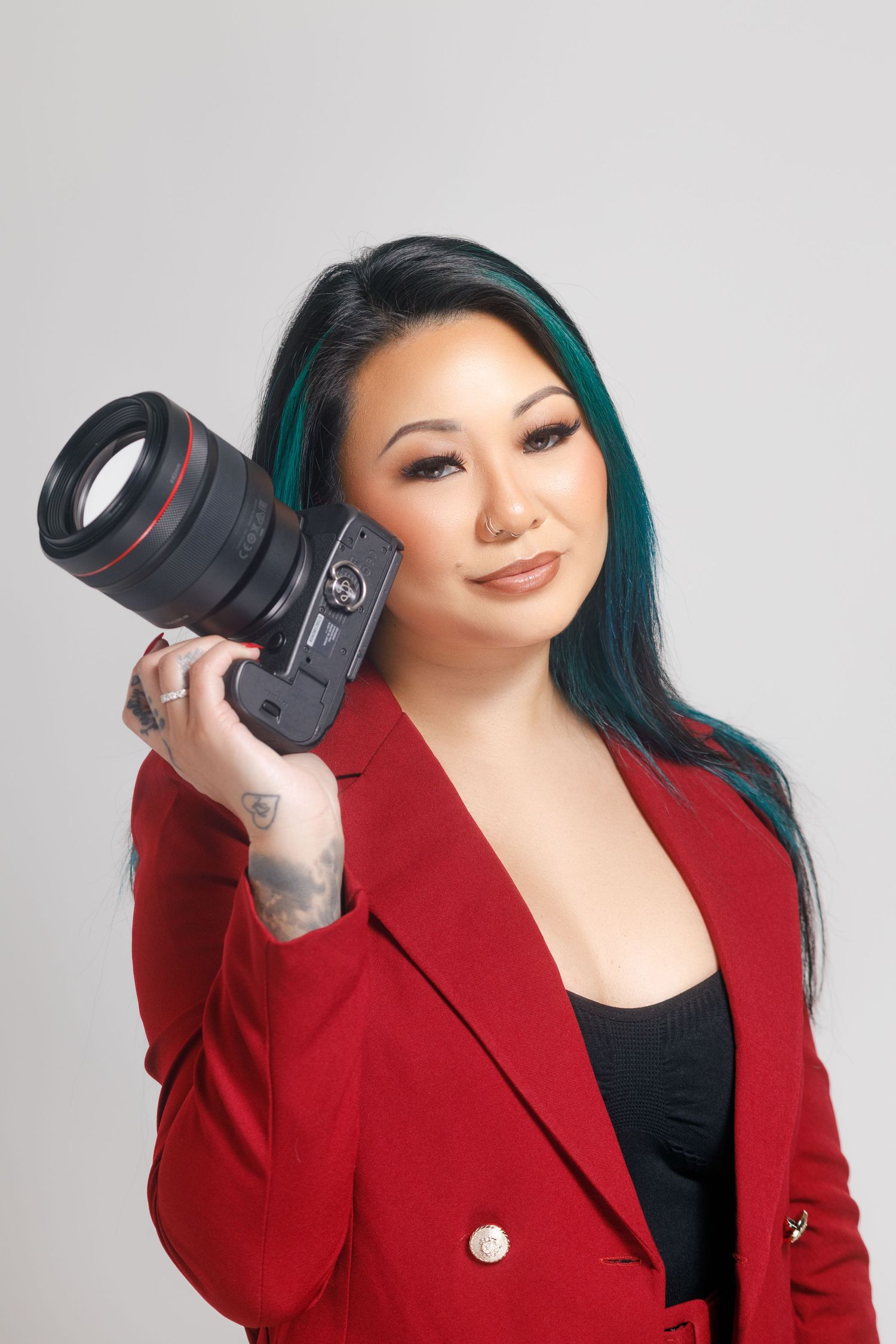 Professional photographer in red blazer and teal hair holds camera while posing against light background.