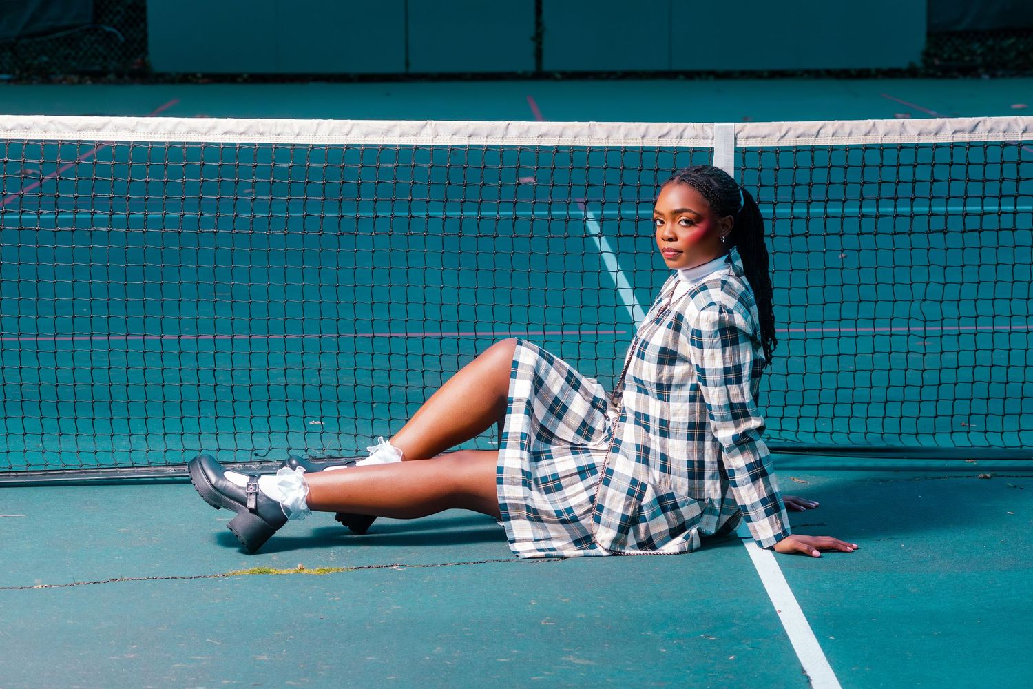 Woman in plaid dress sitting on tennis court by the net, wearing white sneakers in a stylish fashion pose.