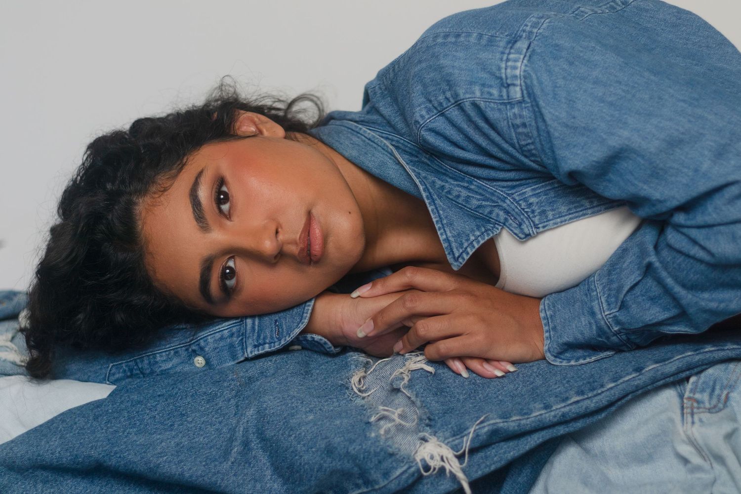 Young woman in denim jacket lying down, gazing at camera with curly hair and subtle makeup against gray background.