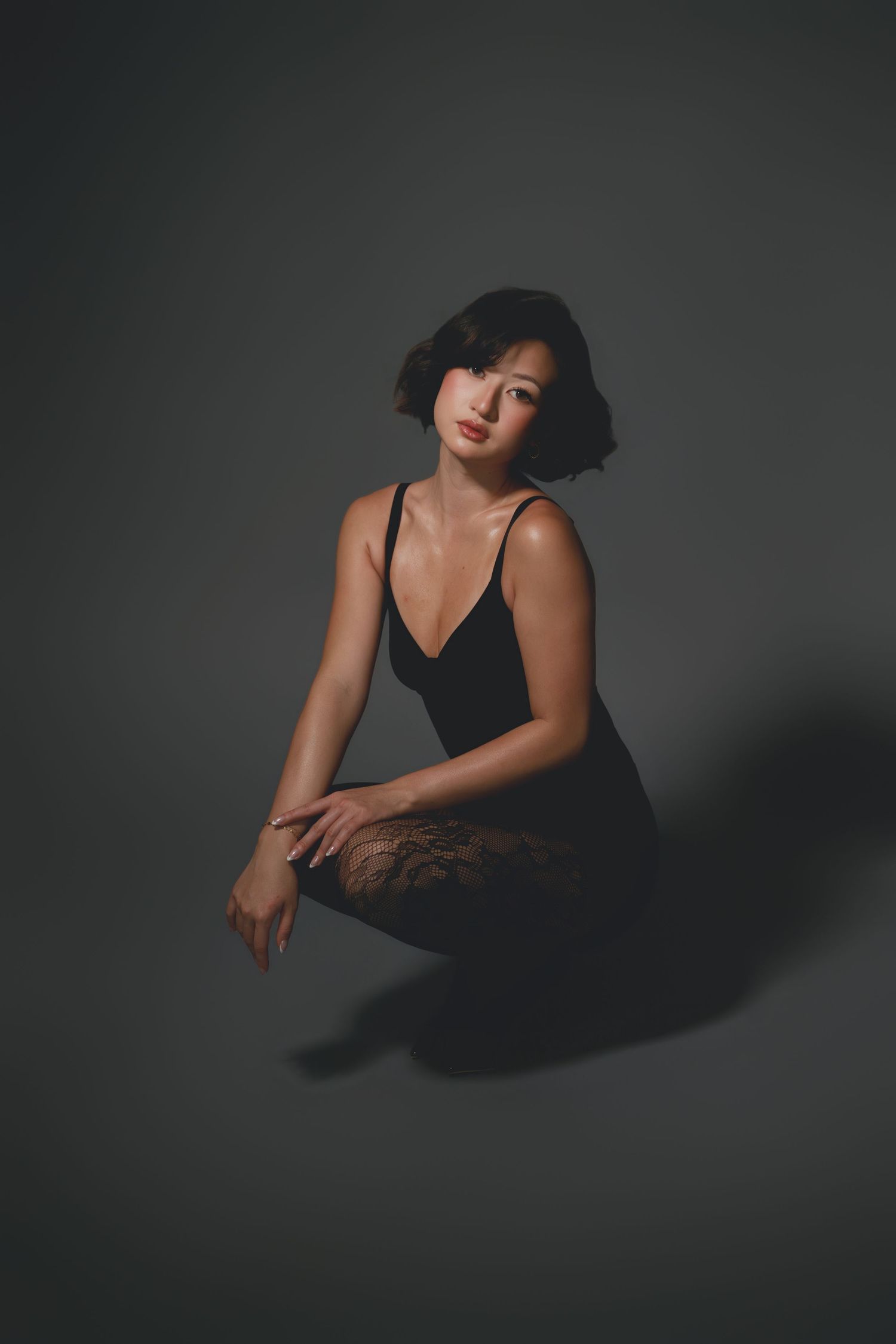 Woman in black bodysuit and lace tights sitting gracefully on floor against dark gray background.