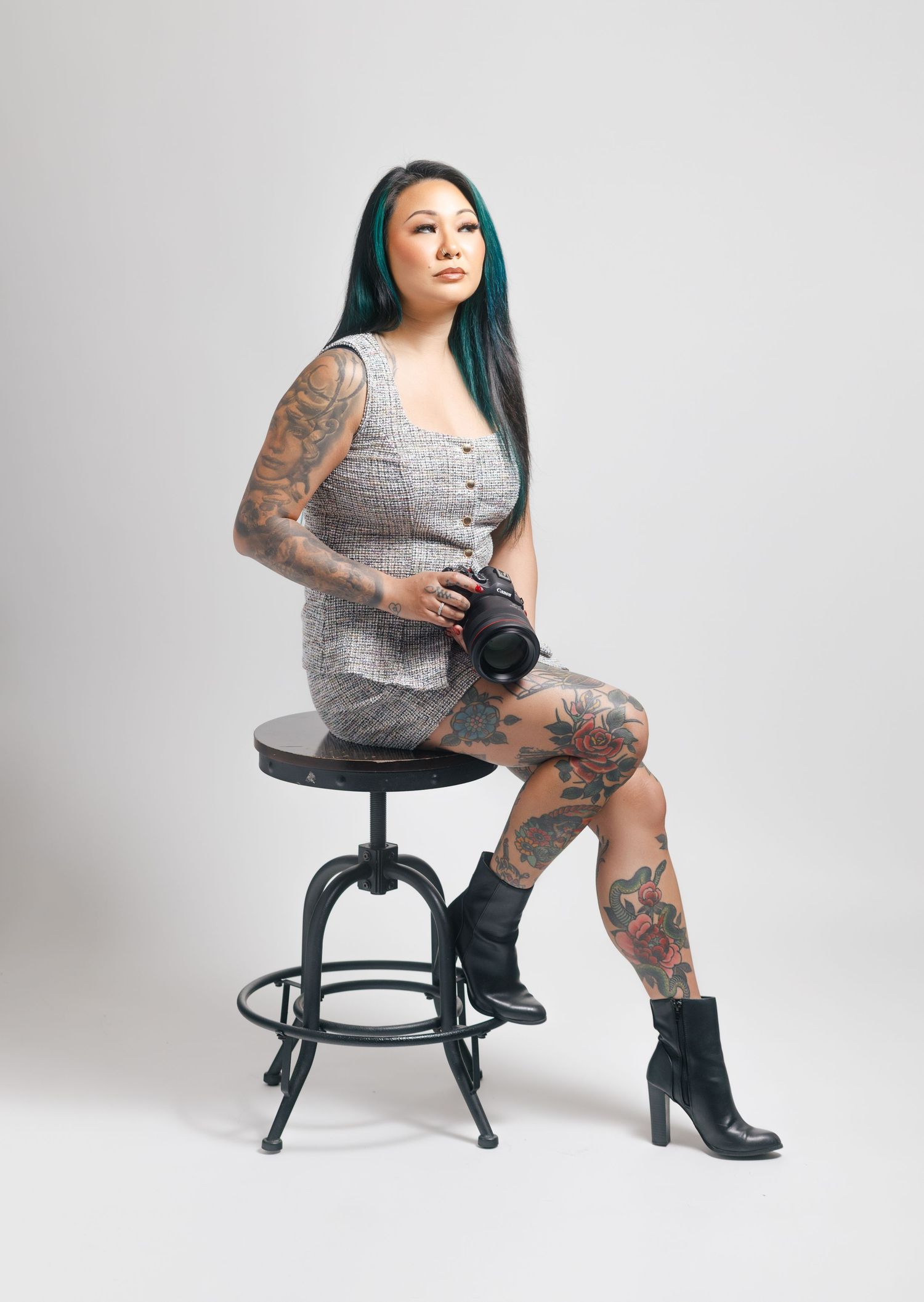Tattooed woman with teal hair sits on stool holding camera, wearing gray dress and black boots.