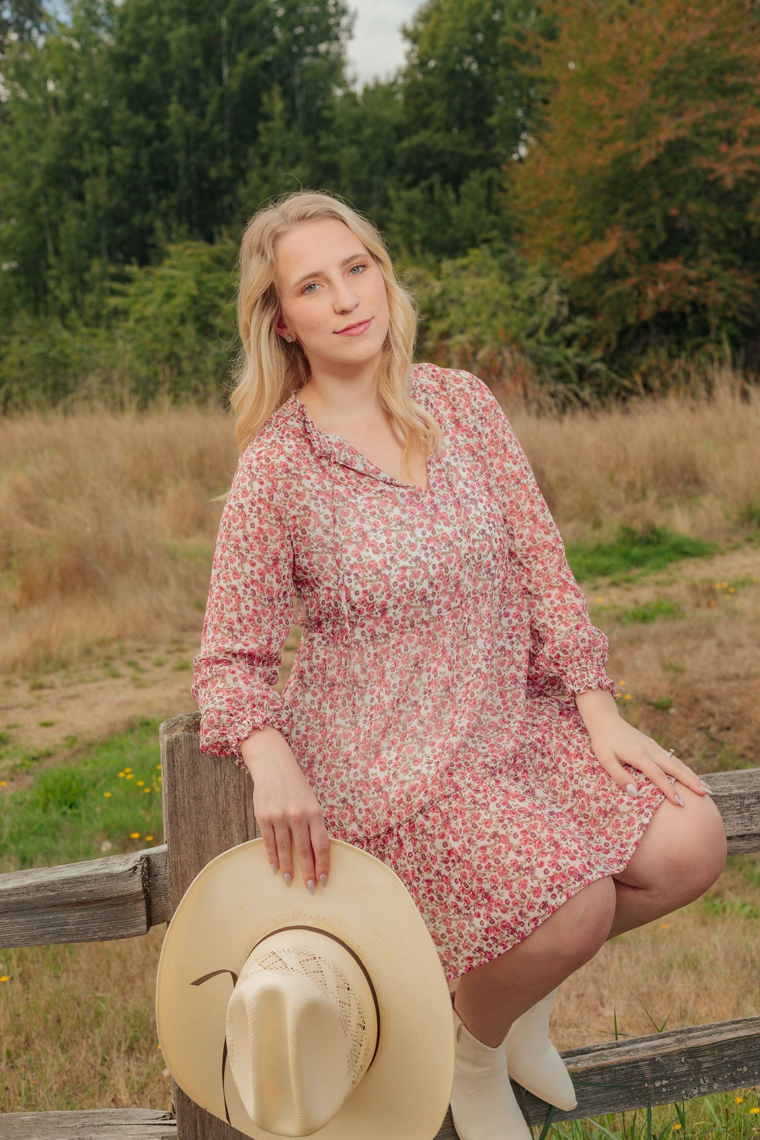 Person in pink floral dress poses with cowboy hat on wooden fence during golden hour.