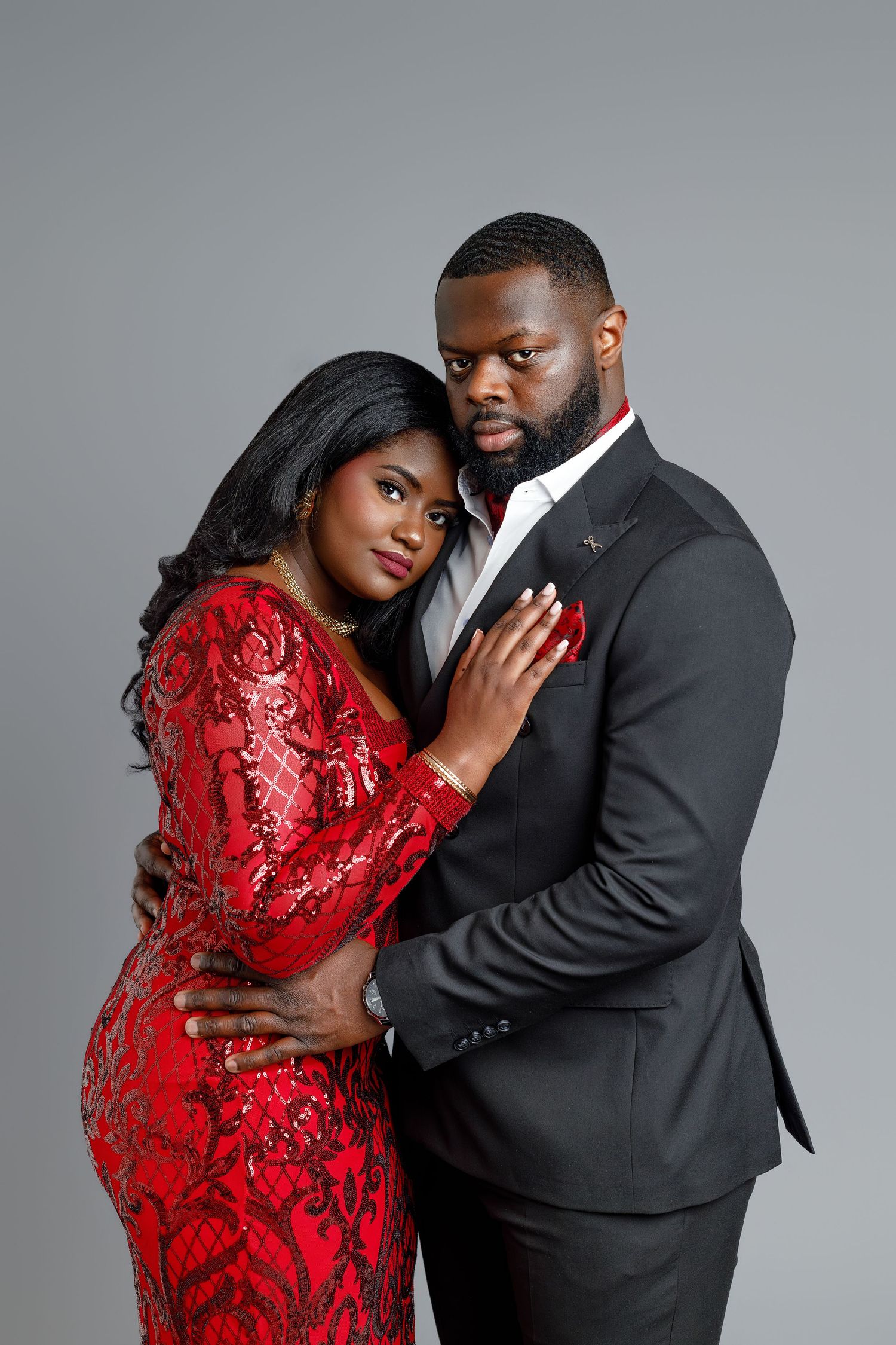 Elegant Black couple posing in studio, woman in red sequin dress, man in black suit, embracing on gray background.