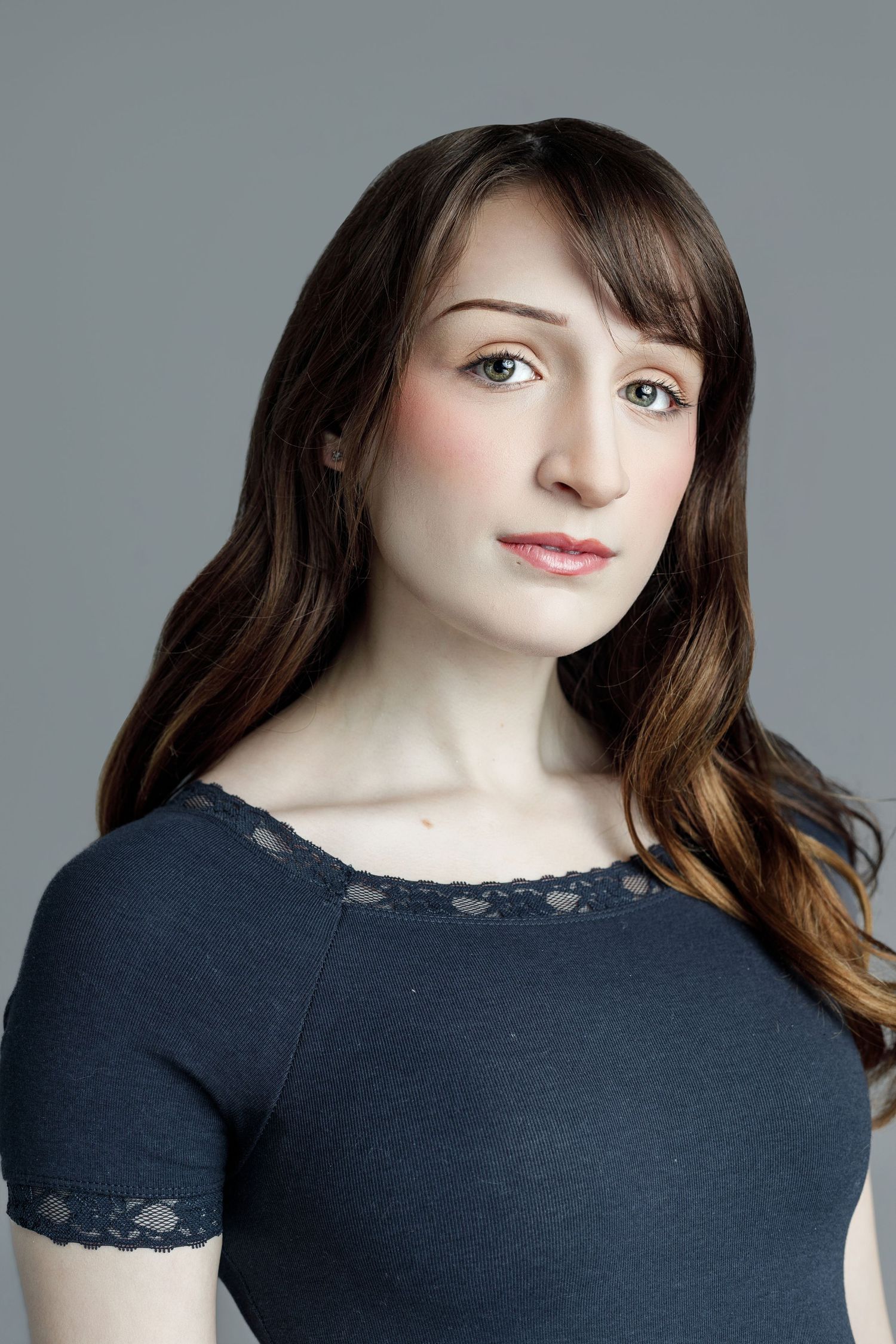 Young woman with long brown hair wearing a dark blue lace-trim top, posing against a gray background.