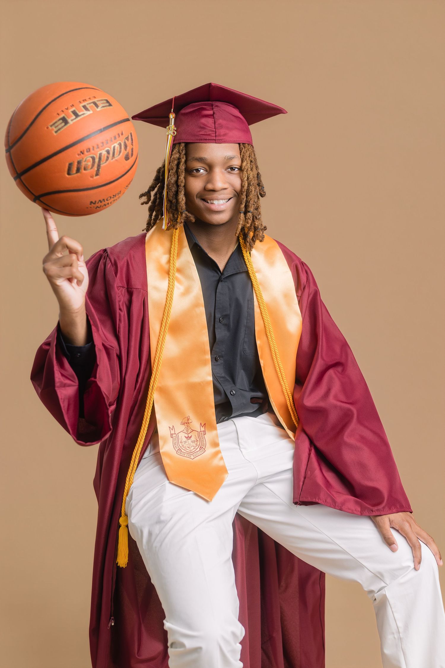 Smiling graduate in maroon cap and gown spinning basketball on finger, wearing gold honor stole and white pants.
