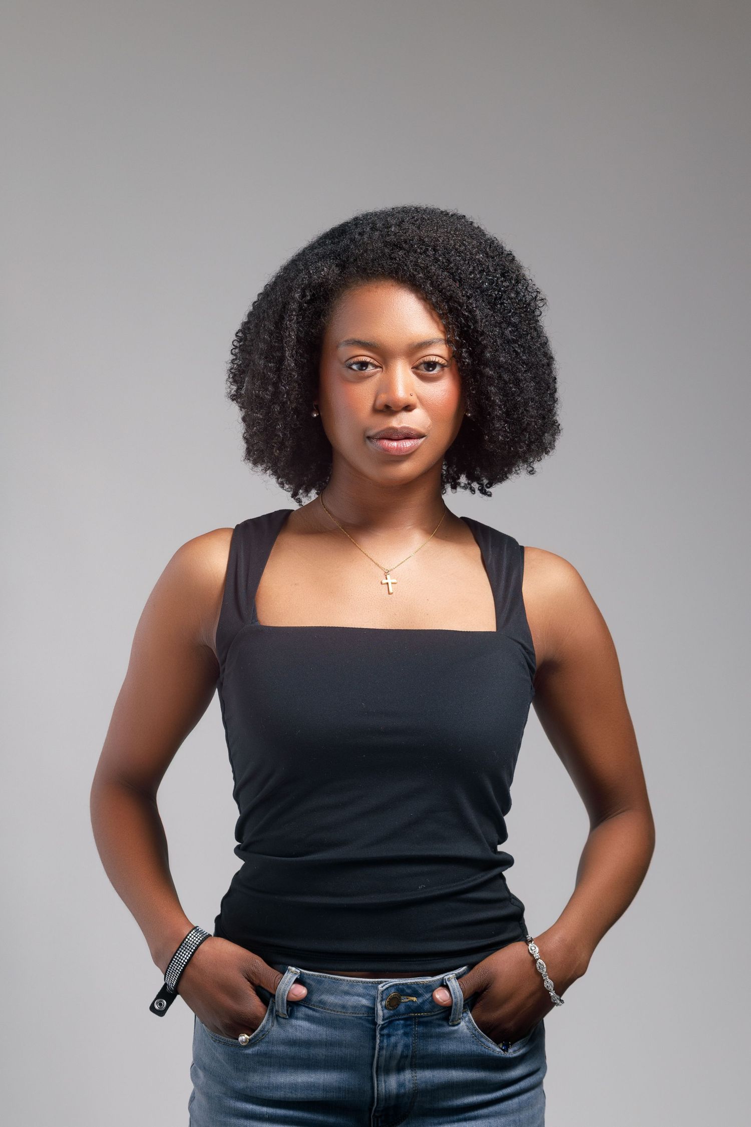 Young Black woman with natural hair in black tank top and jeans, hands in pockets, wearing gold cross necklace.