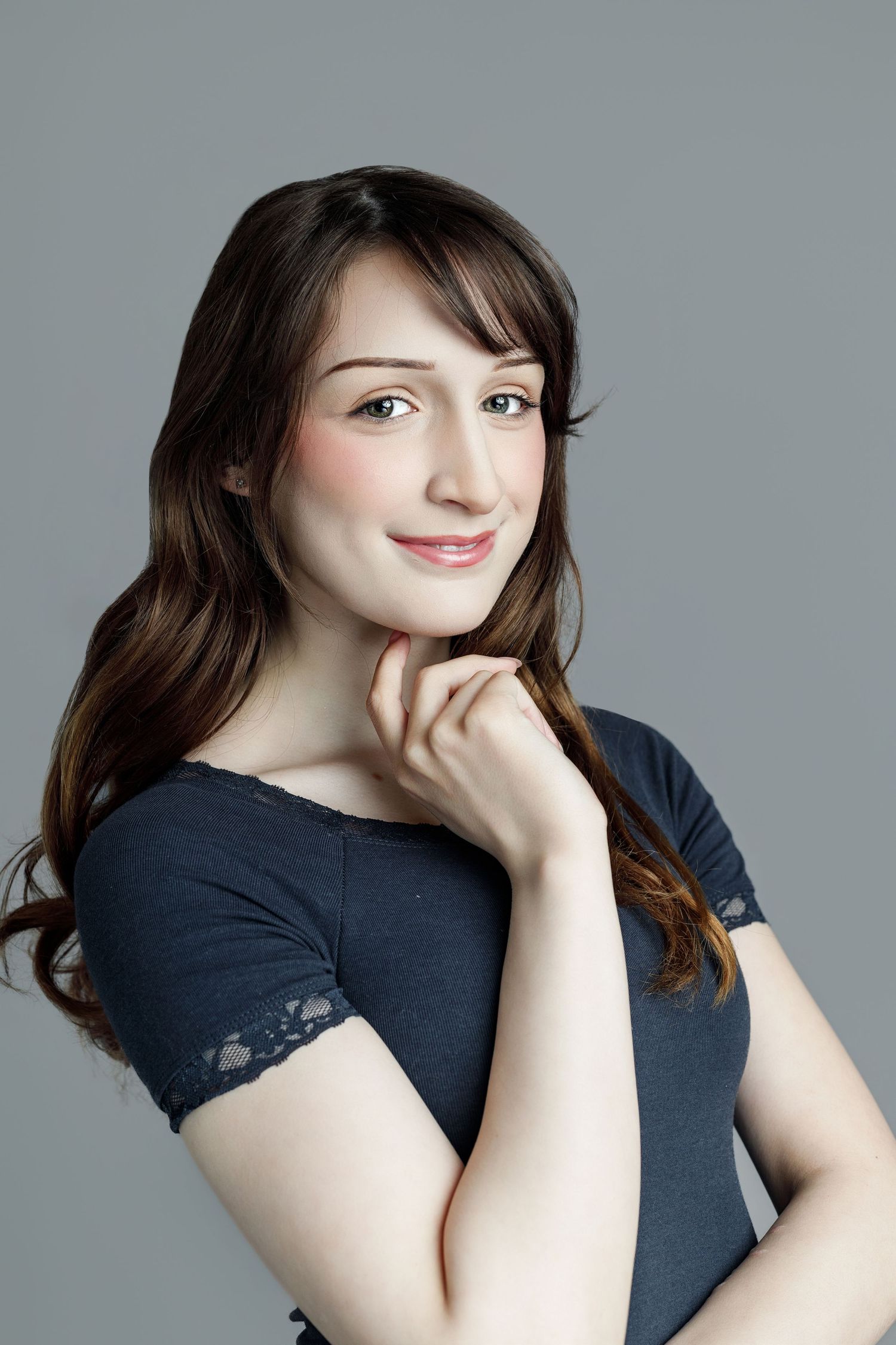 Young woman with long brown hair smiling softly, wearing a gray top, posing against a gray background.
