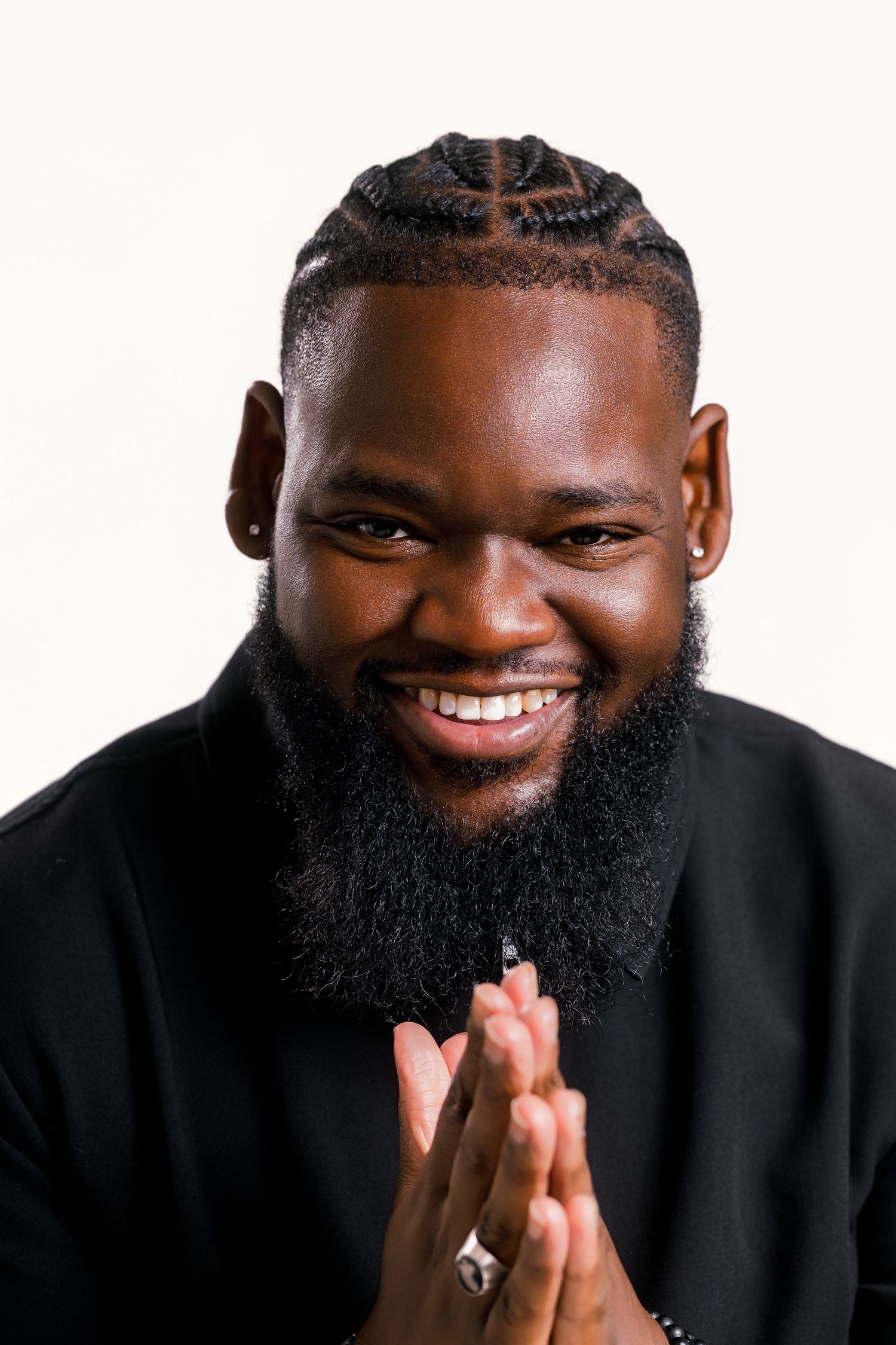 Smiling Black man with braided hair and beard, wearing black, hands clasped, posing against white background.