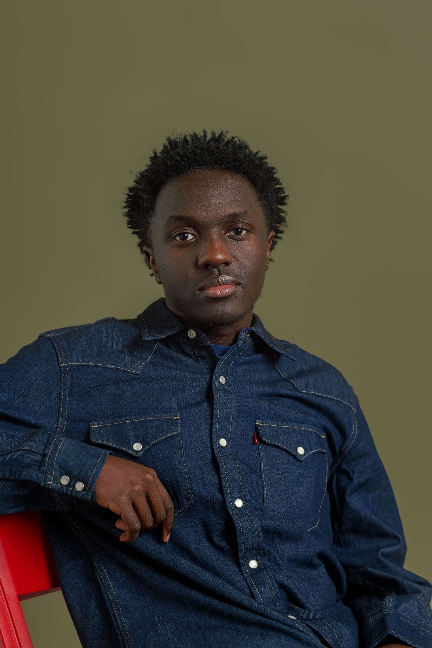Young Black man in a dark denim Levi's shirt sits relaxed on a red chair against an olive green background.