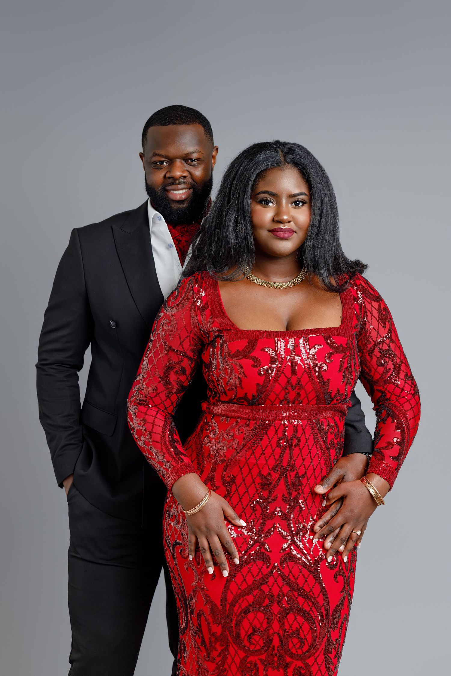 Elegant couple posing in studio, man in black suit, woman in red sequin gown, gray background.
