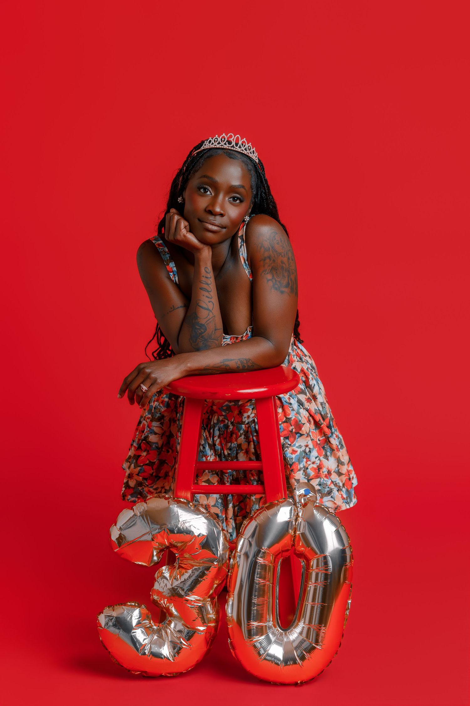 Woman in floral dress with tiara poses by red stool with 30 balloons, celebrating 30th birthday on red background.