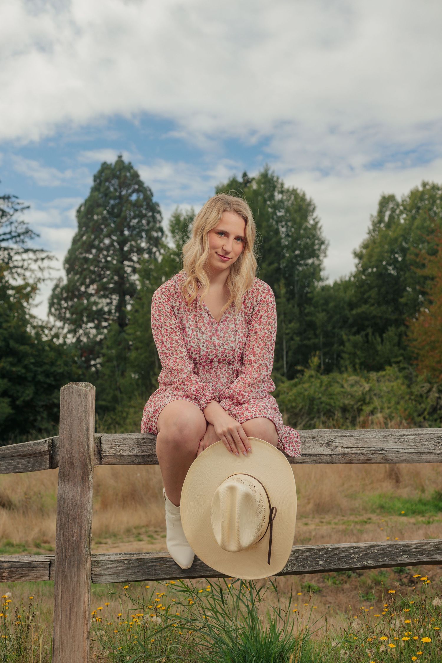 Person sitting on a wooden fence wearing a pink floral dress and holding a beige cowboy hat.