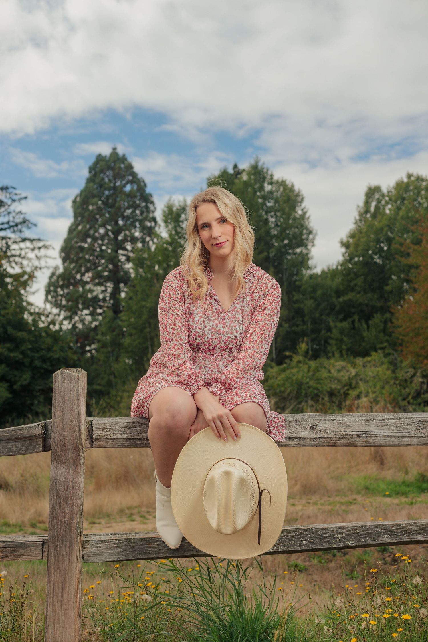 Blonde woman in floral dress and white boots sitting on rustic wooden fence holding cowboy hat with trees behind her.