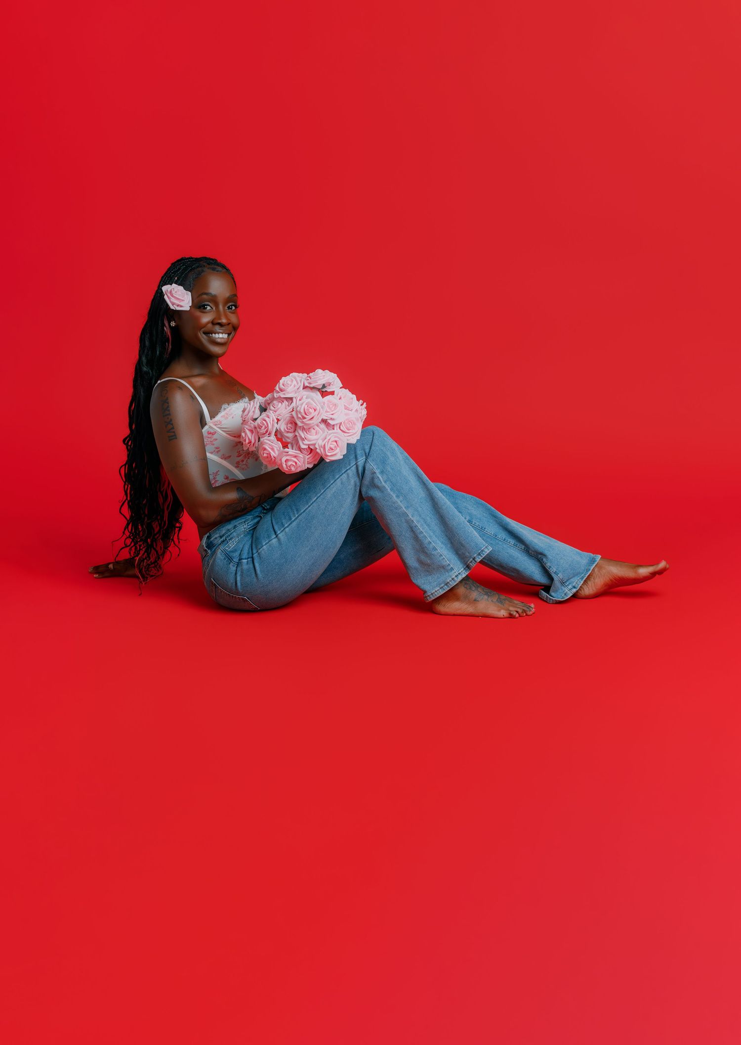 Smiling woman sitting on red background holding pink flowers, wearing jeans and white top with flower hair accessory.